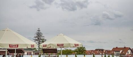 Un espace extérieur aménagé avec des tables et des chaises sous des parasols blancs, entouré de plantes en pot et d’une pelouse verdoyante.