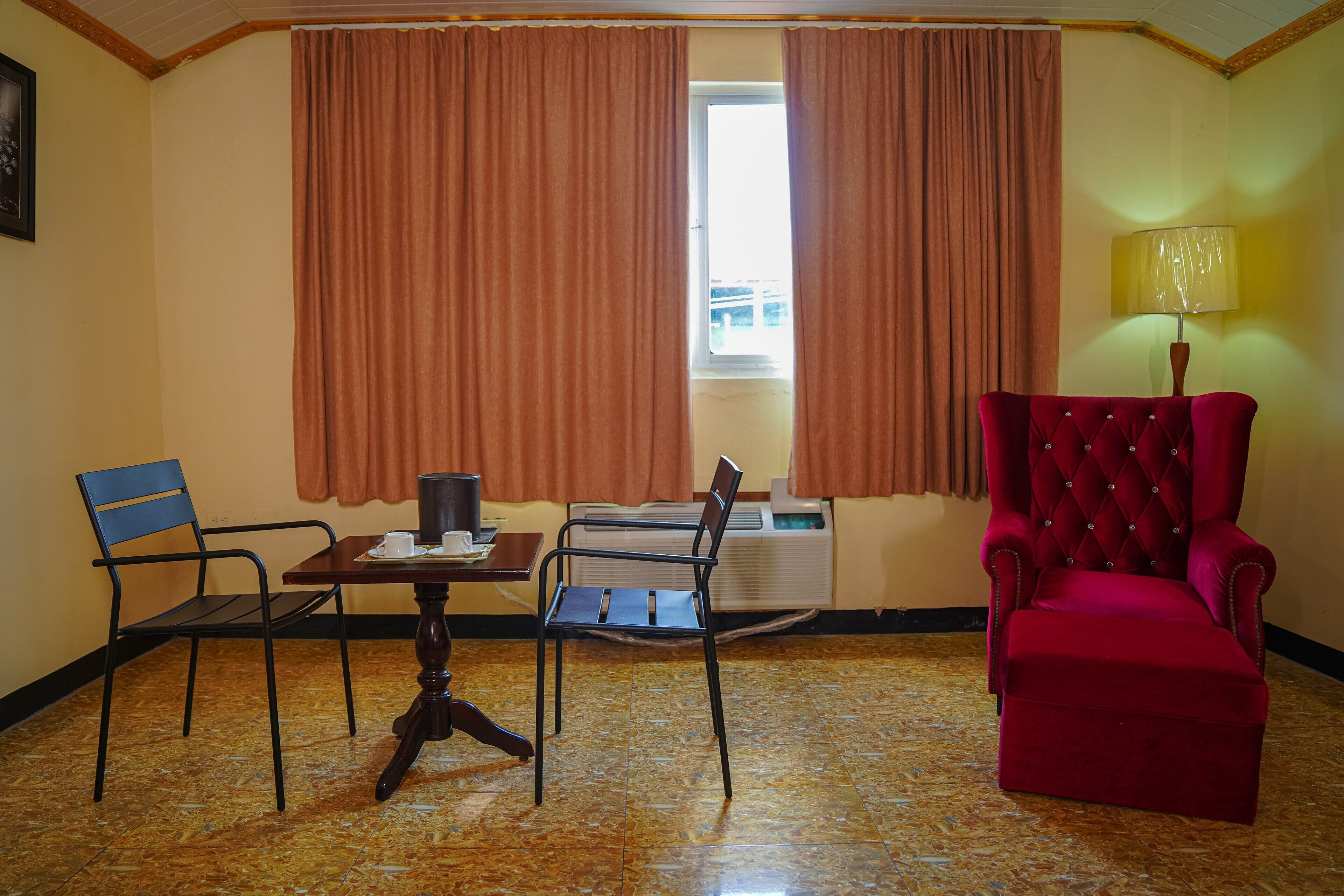 A room with a dining table, two chairs, a red armchair, a lamp, and a window with curtains.
