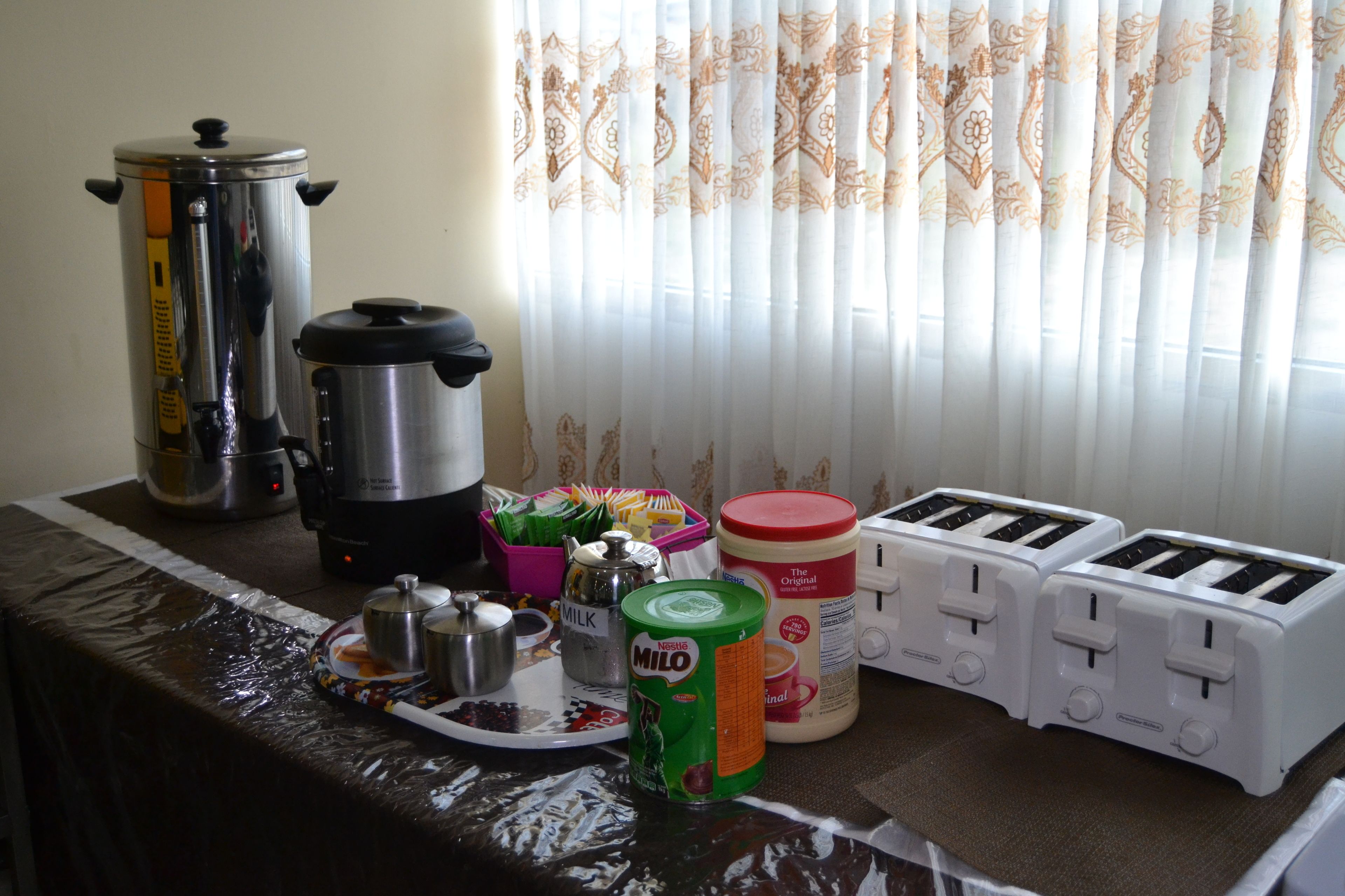 A kitchen counter with a coffee maker, a rice cooker, a milk container, a can of Milo, and two toasters.