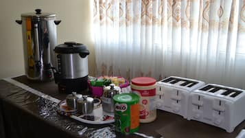 Una encimera de cocina con una cafetera, una cocedora de arroz, un depósito de leche, una lata de Milo y dos tostadoras.