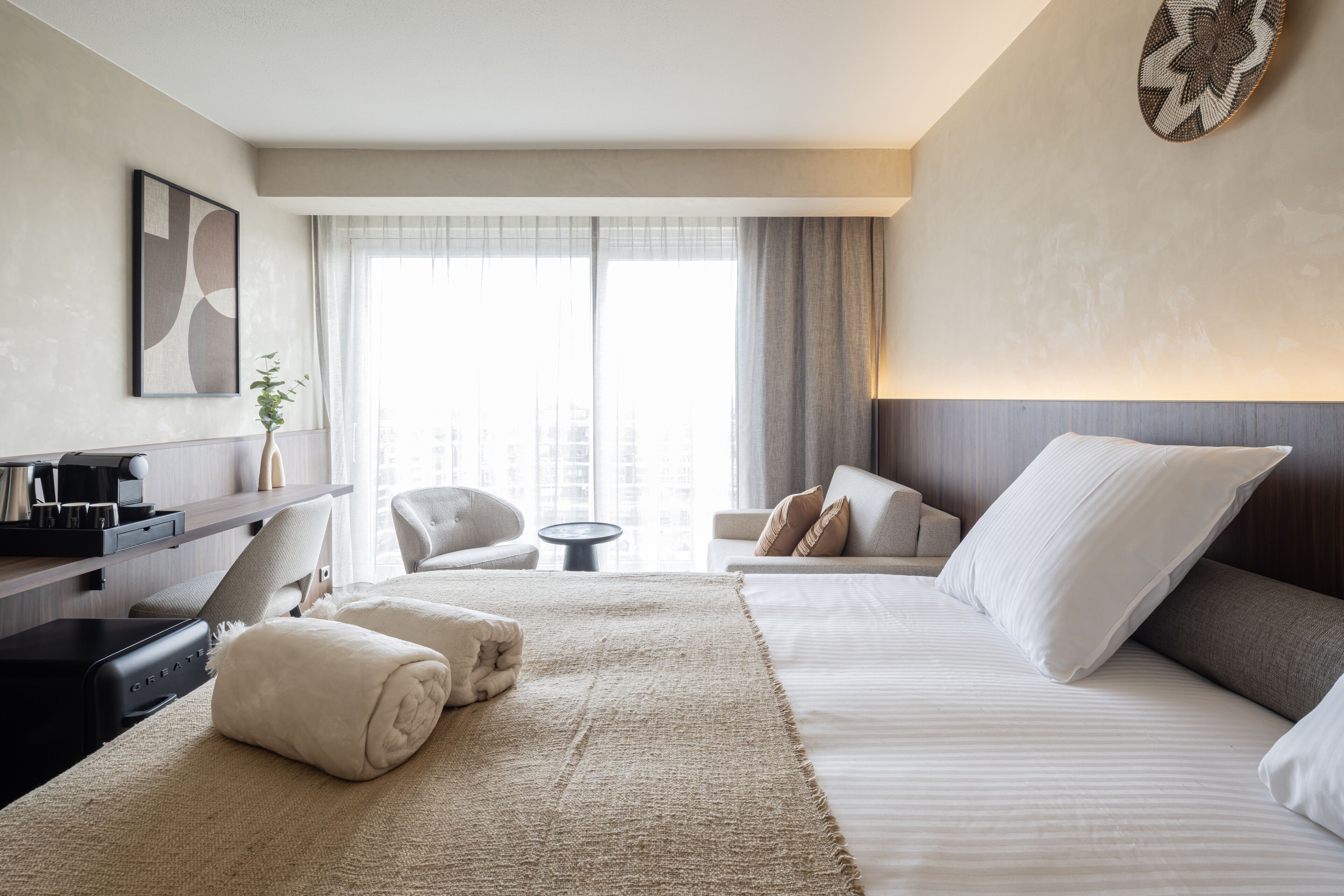Une chambre d’hôtel moderne avec un grand lit, un bureau et une chaise. On y trouve une fenêtre avec des rideaux, une œuvre d’art accrochée au mur et une petite table avec une plante.