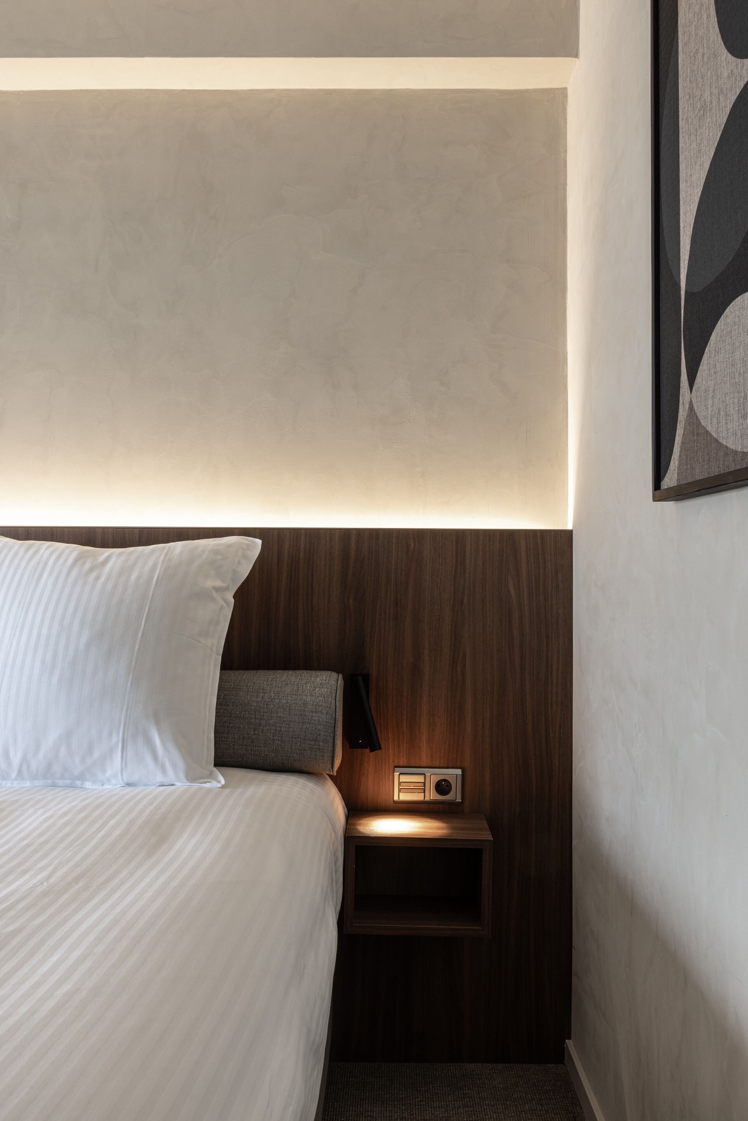 A hotel room with a neatly made bed, a bedside table with a radio, and a wall-mounted artwork.