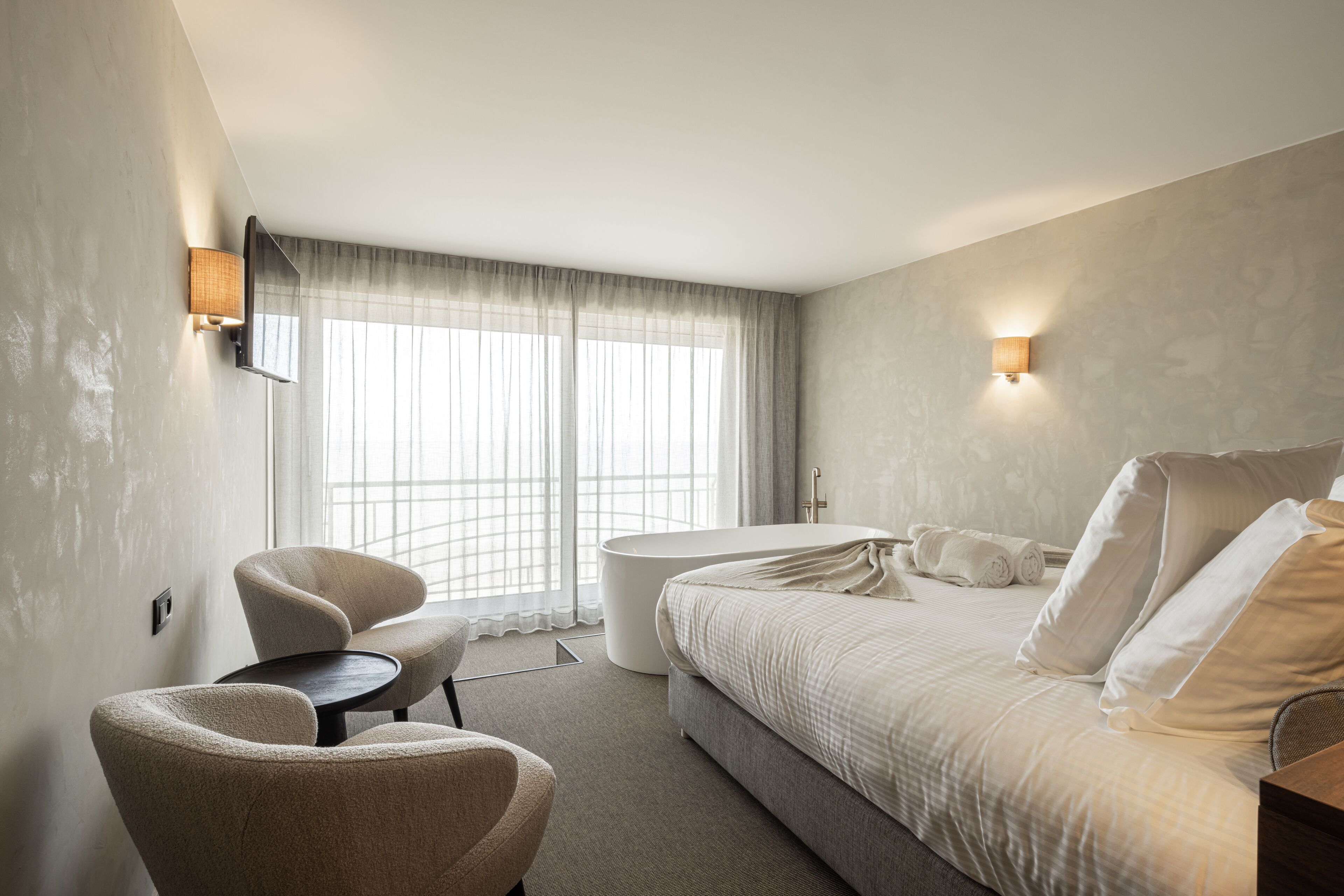 Una habitación de hotel moderna con una cama grande, una mesa de centro redonda, dos sillones, una mesita y un ventanal con cortinas translúcidas.