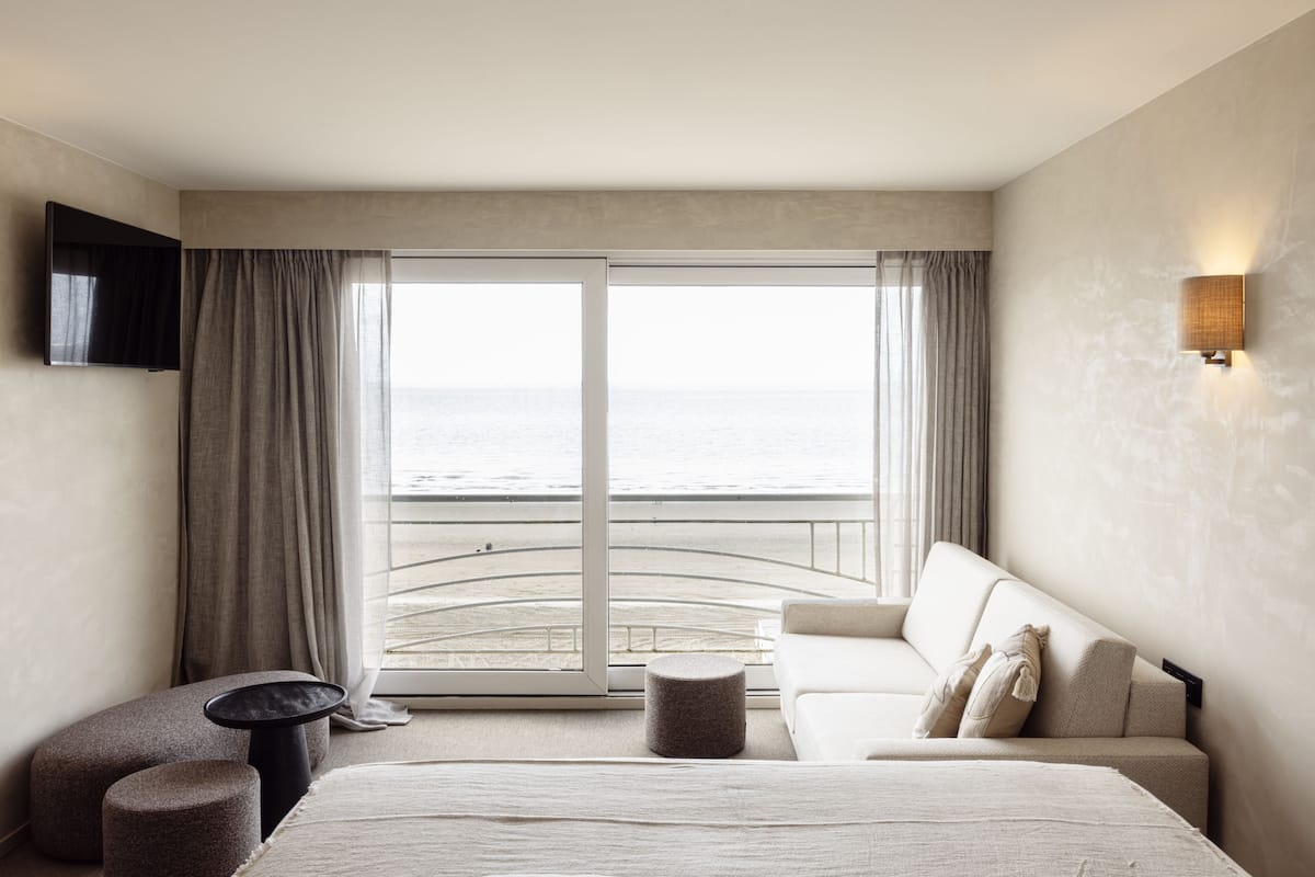 Habitación de hotel con una cama grande, un sofá, una mesita y un televisor instalado en la pared. La habitación tiene vista a la playa y al océano a través de amplios ventanales.