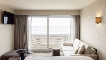 Een hotelkamer met een groot bed, een bank, een kleine tafel en een aan de muur bevestigde televisie. De kamer heeft uitzicht op het strand en de oceaan door grote ramen.