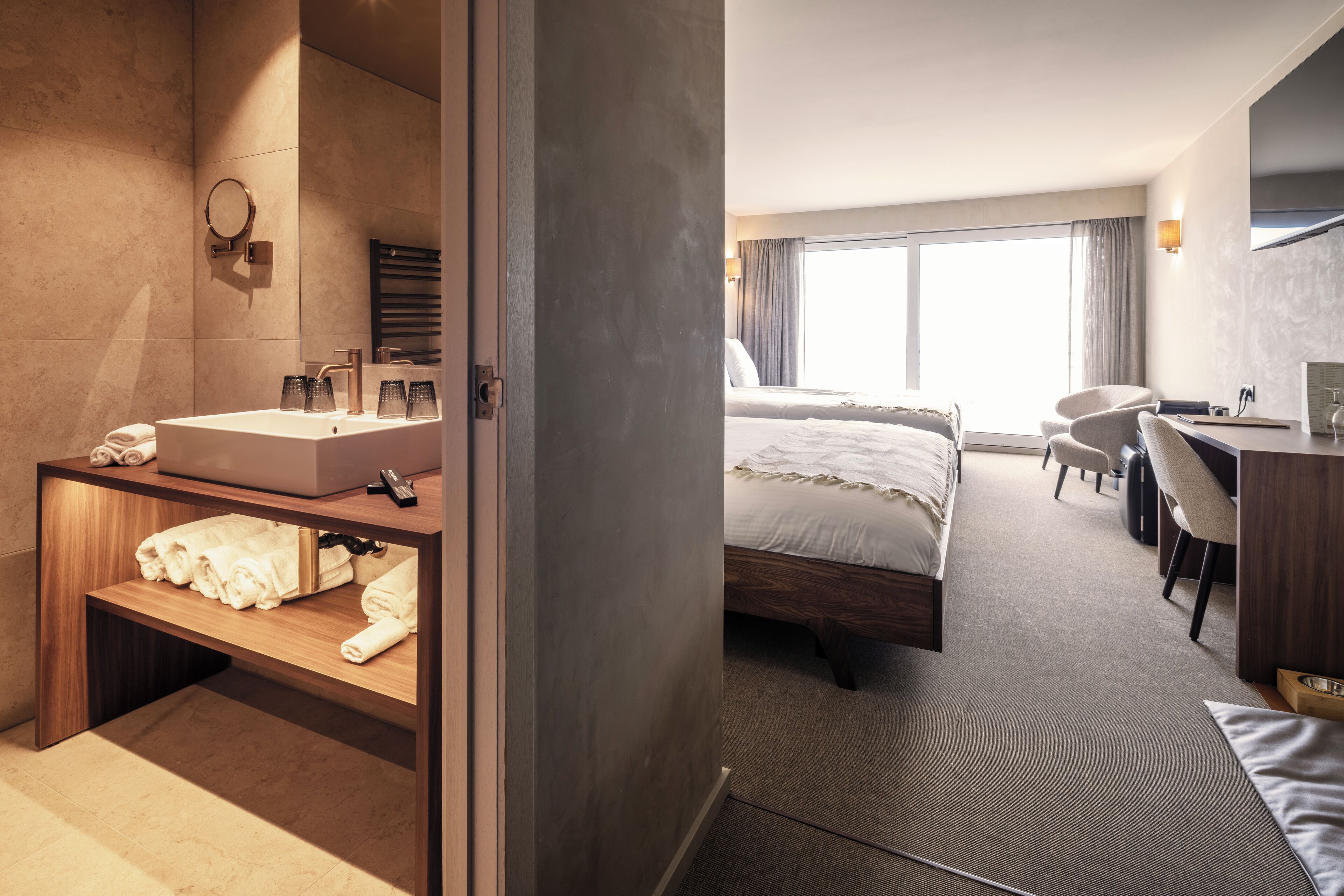 Une chambre d’hôtel moderne équipée d’un grand lit, d’un bureau, d’une chaise, d’une coiffeuse avec un miroir et d’une salle de bain avec un lavabo et un porte-serviettes.