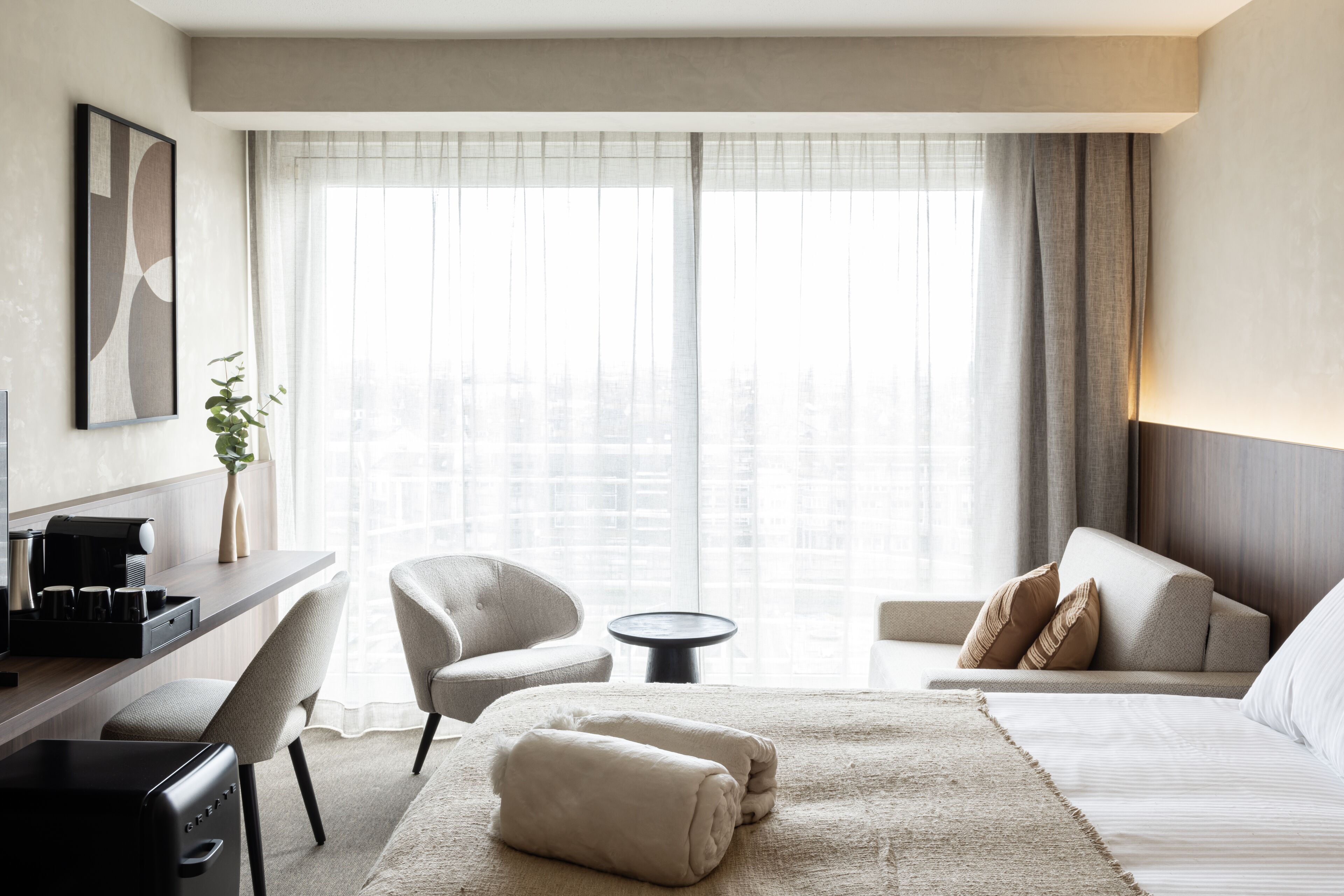 A hotel room with a bed, a sofa, a desk with a coffee maker, a chair, and a view of a cityscape through large windows.