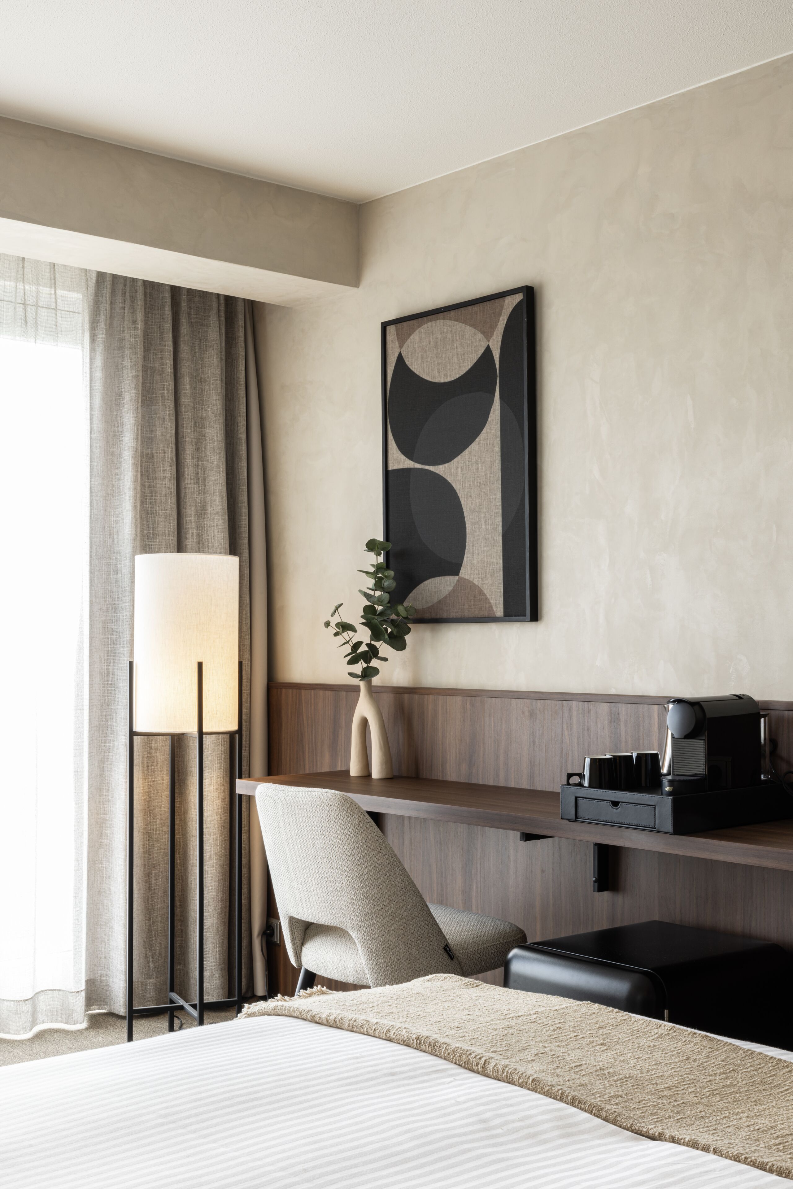 A hotel room with a bed, a desk, a lamp, a chair, a plant, and a coffee machine.