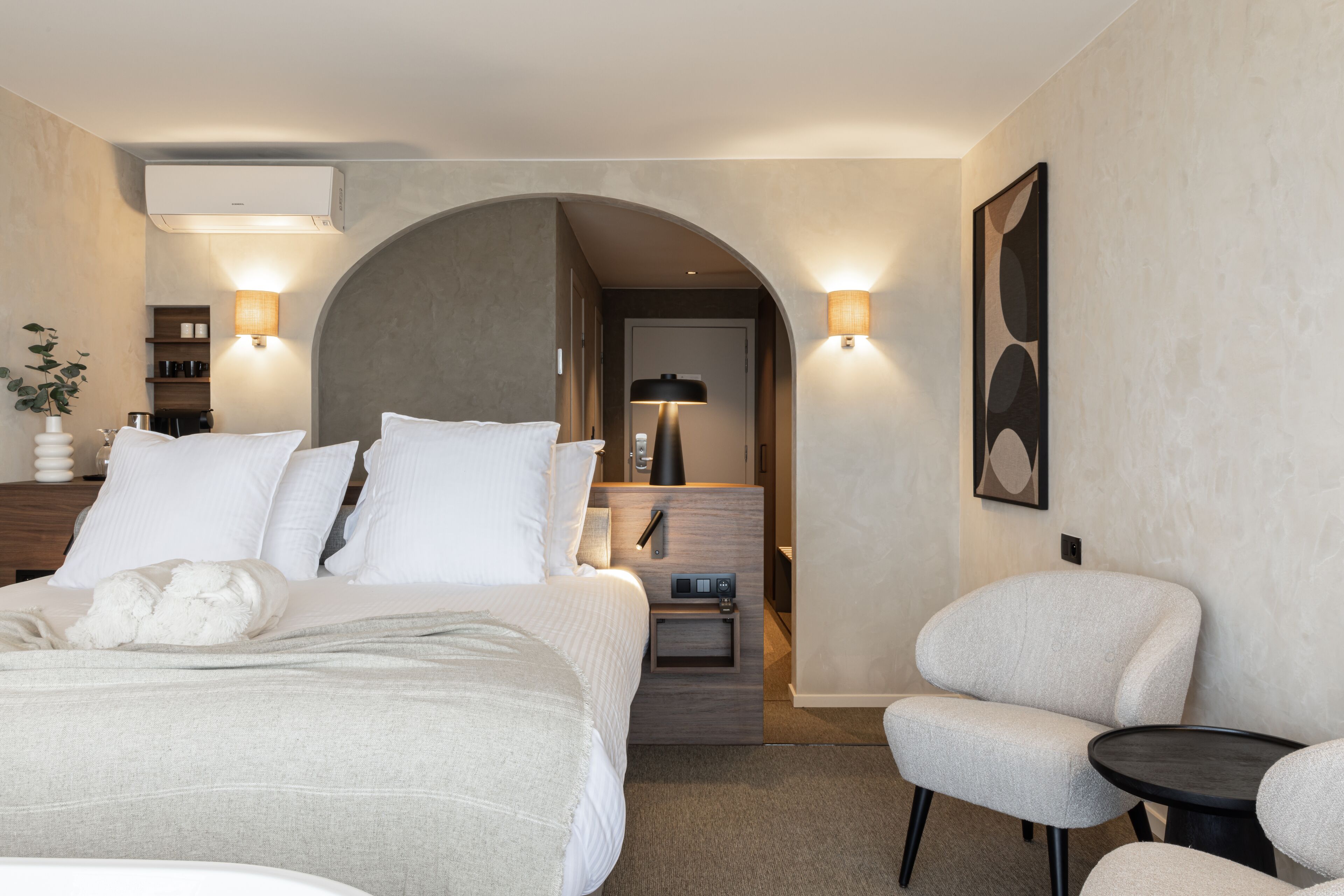 Une chambre d’hôtel moderne dotée d’un grand lit, d’un coin salon avec un fauteuil et une petite table, et d’un couloir menant à la salle de bain.