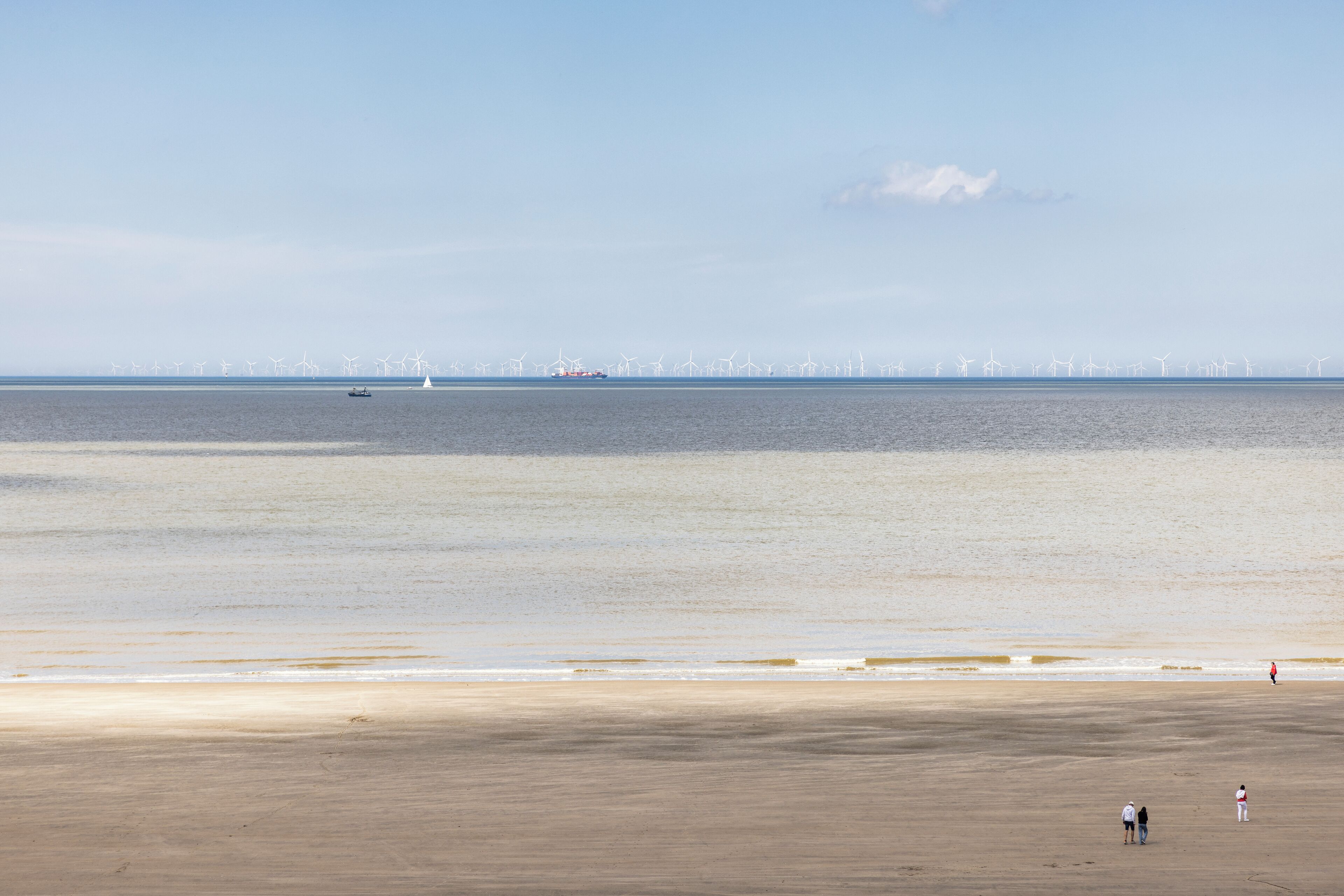 Une plage avec une mer calme, des éoliennes visibles au loin et quelques personnes se promenant sur le rivage.