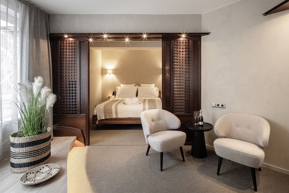 Una habitación de hotel moderna con una cama, dos sillones, una mesita, una planta en maceta y un panel decorativo en la pared.