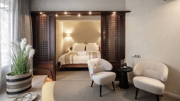 A modern hotel room with a bed, two armchairs, a small table, a potted plant, and a decorative wall panel.