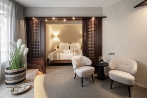 A modern hotel room with a bed, two armchairs, a small table, a potted plant, and a decorative wall panel.