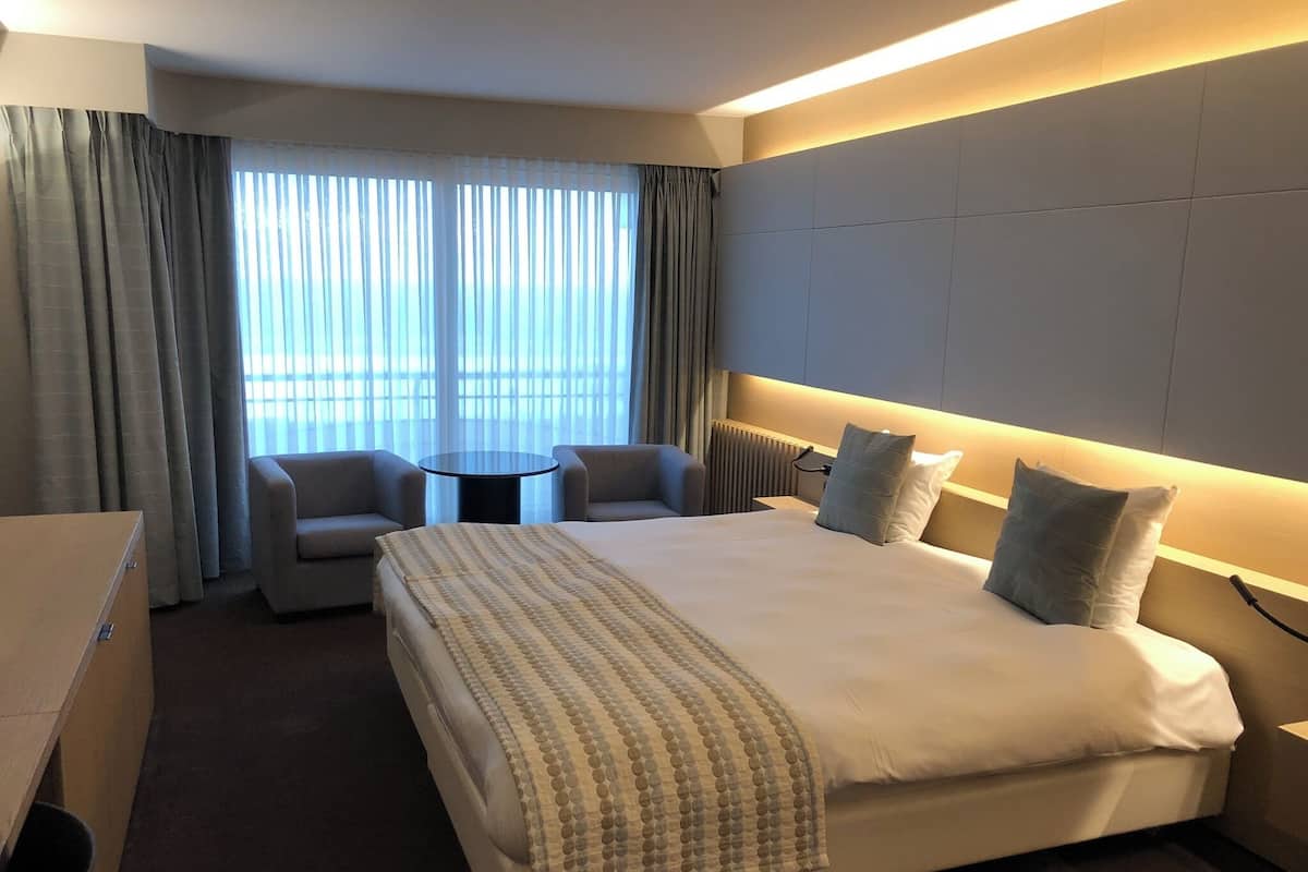 Habitación de hotel con una cama grande, dos sillones, una mesita y vistas a la ciudad a través de amplias ventanas.