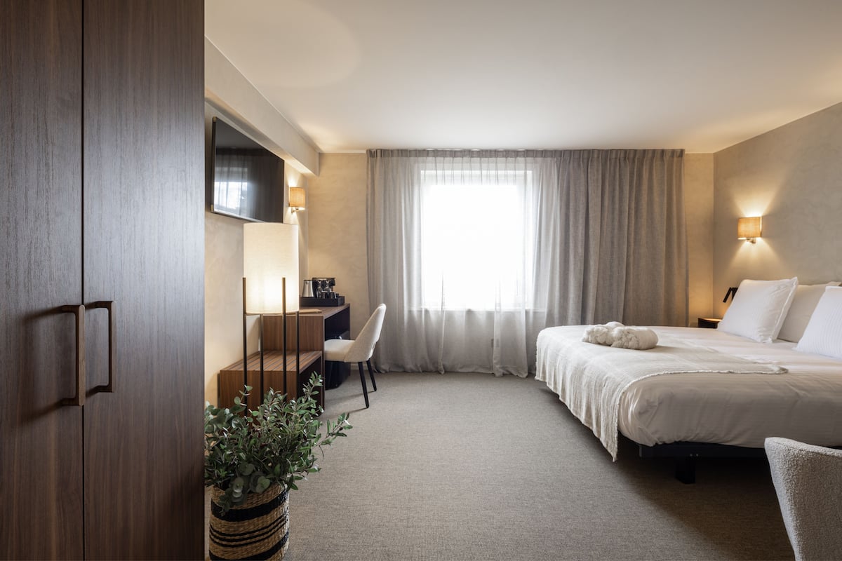 Una habitación de hotel con cama, escritorio con silla, televisor y ventana con cortinas.