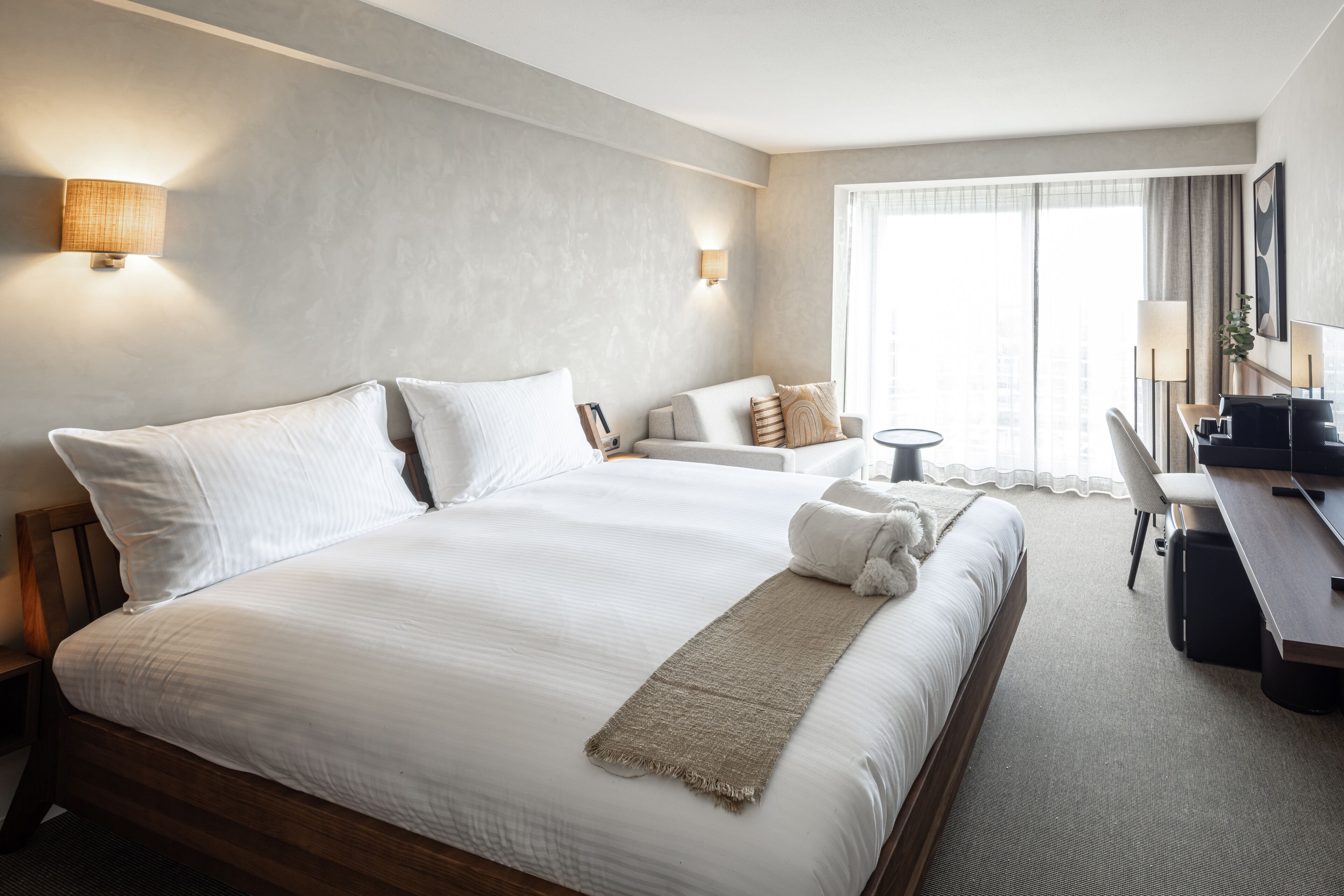 Habitación de hotel con una cama grande, un escritorio y una silla. Hay dos lámparas de pared y una ventana con cortinas.