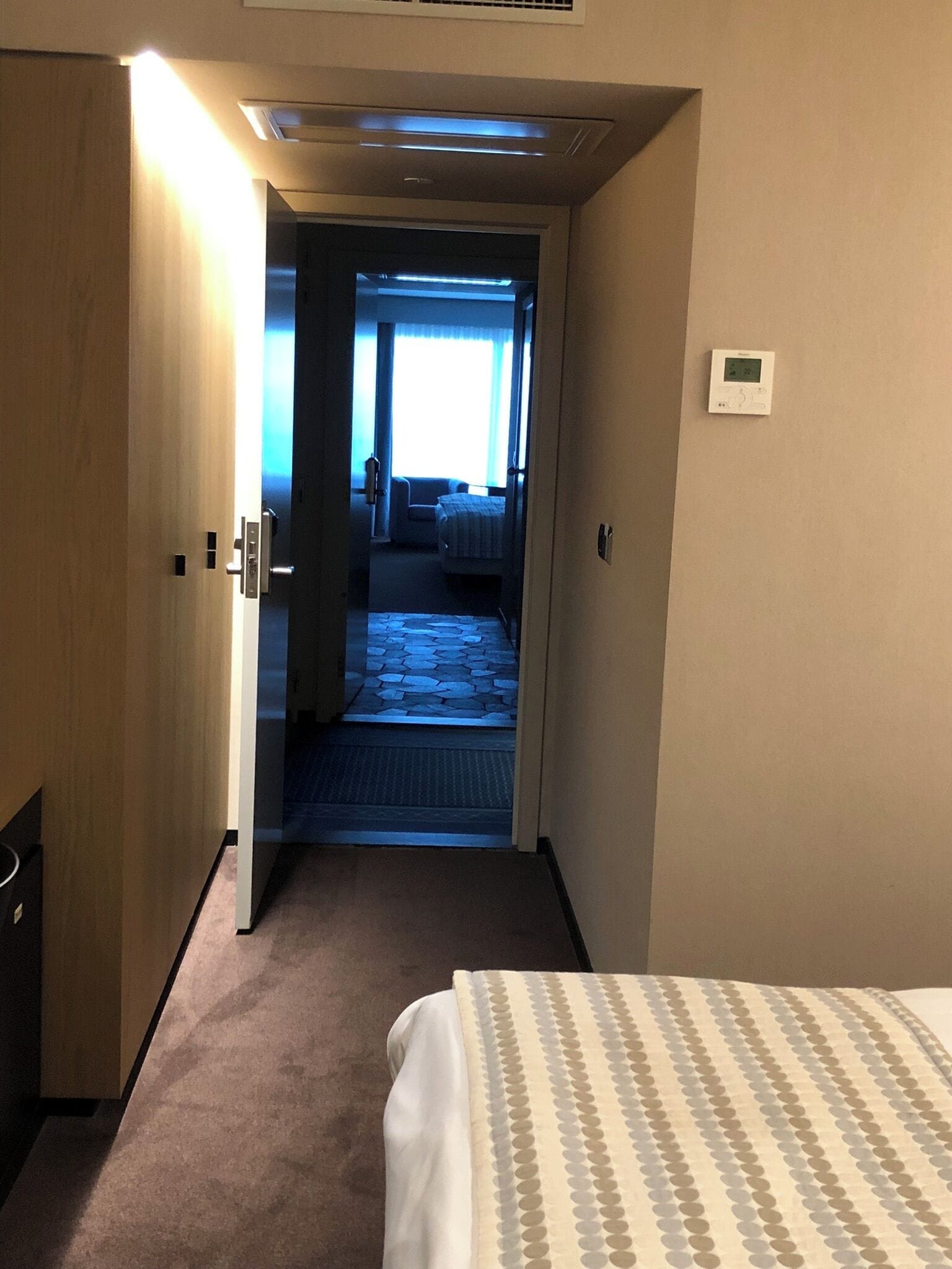 Habitación de hotel con cama, puerta, termostato y vista a otra habitación con cama y ventana.