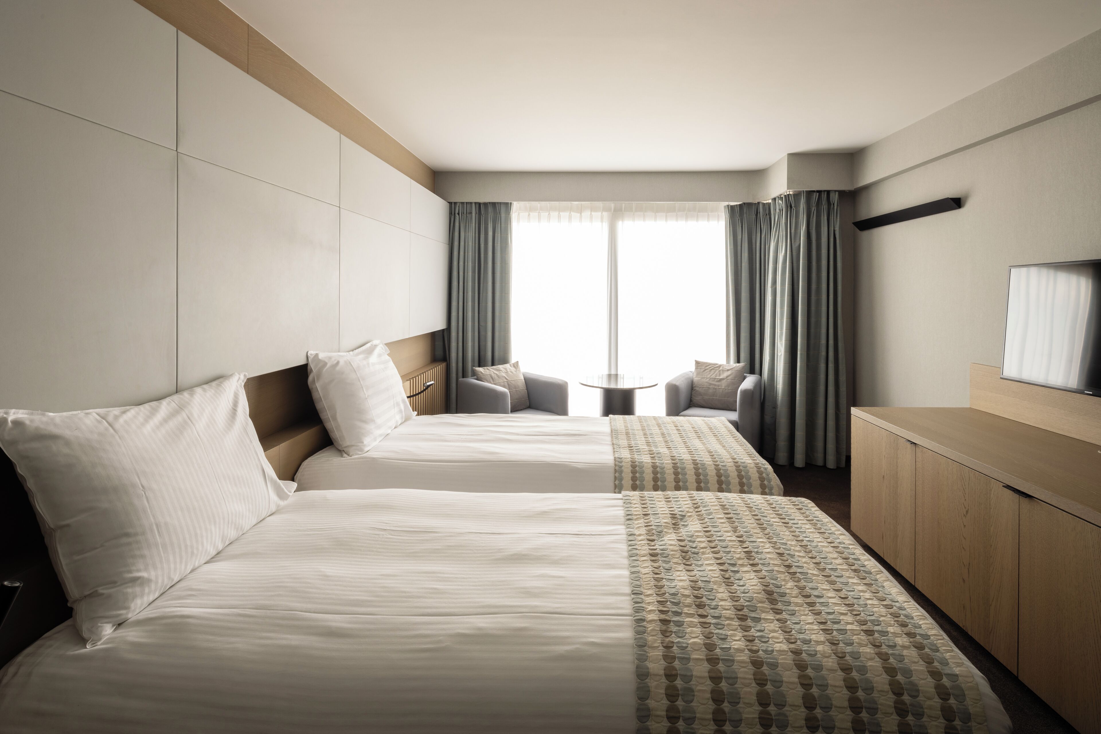 Quarto de hotel com duas camas, uma mesa redonda pequena, uma televisão e vista para o exterior.