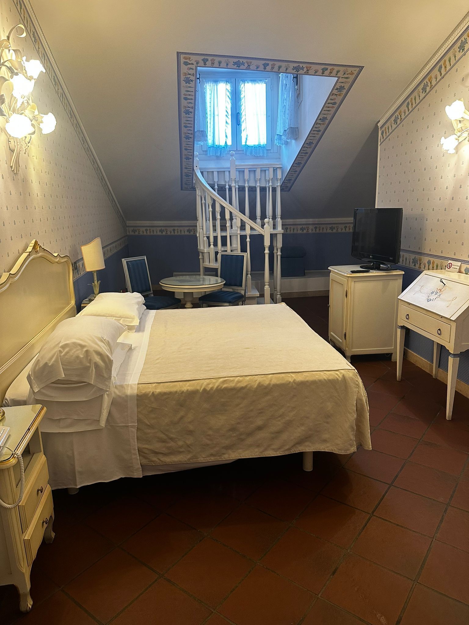 Un dormitorio con cama, mesita de noche, lámpara, escritorio, silla y televisor.