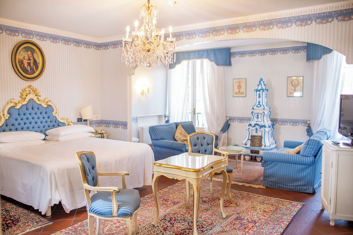 A hotel room with a large bed, a sitting area with a table and chairs, a chandelier, and a decorative cabinet.