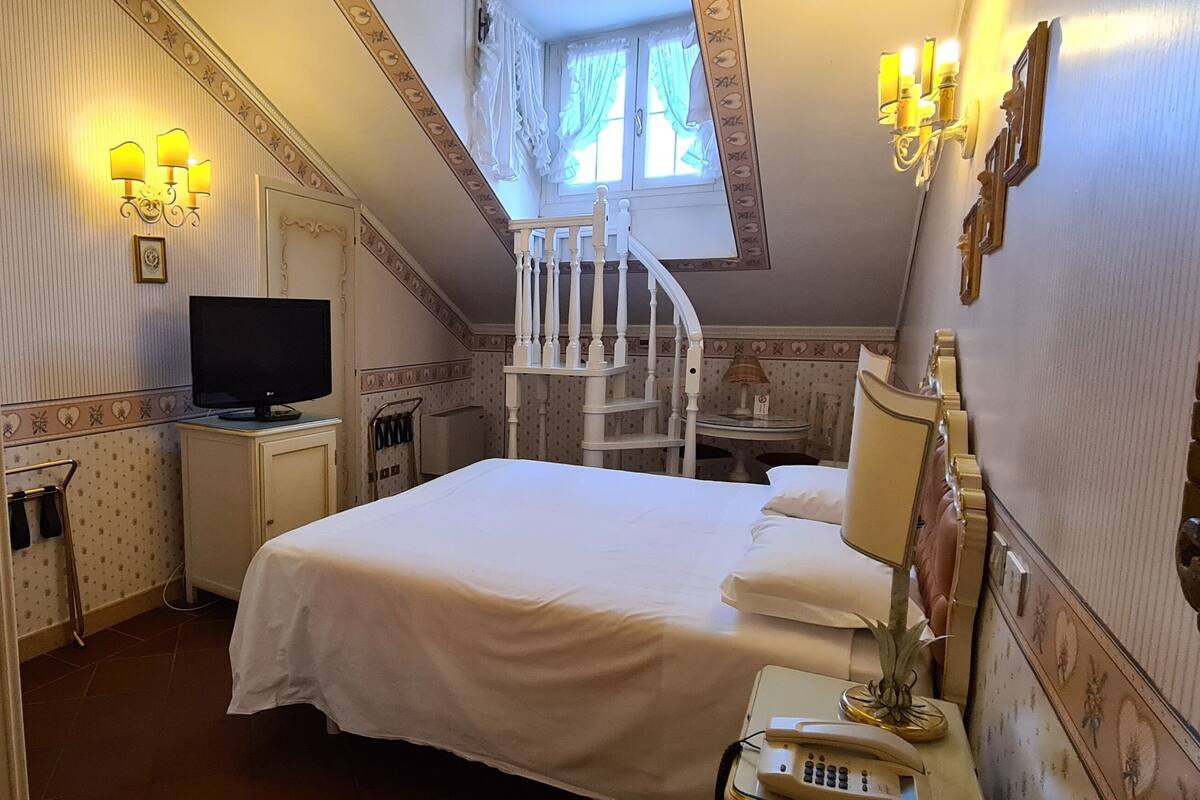 A bedroom with a bed, a television, a small table with a telephone, a mirror, and a staircase leading to an upper level.