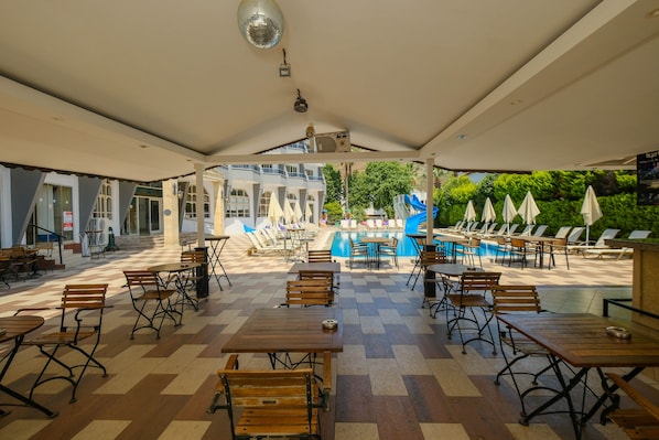 Un área de patio exterior con mesas y sillas de madera, una piscina y sombrillas.