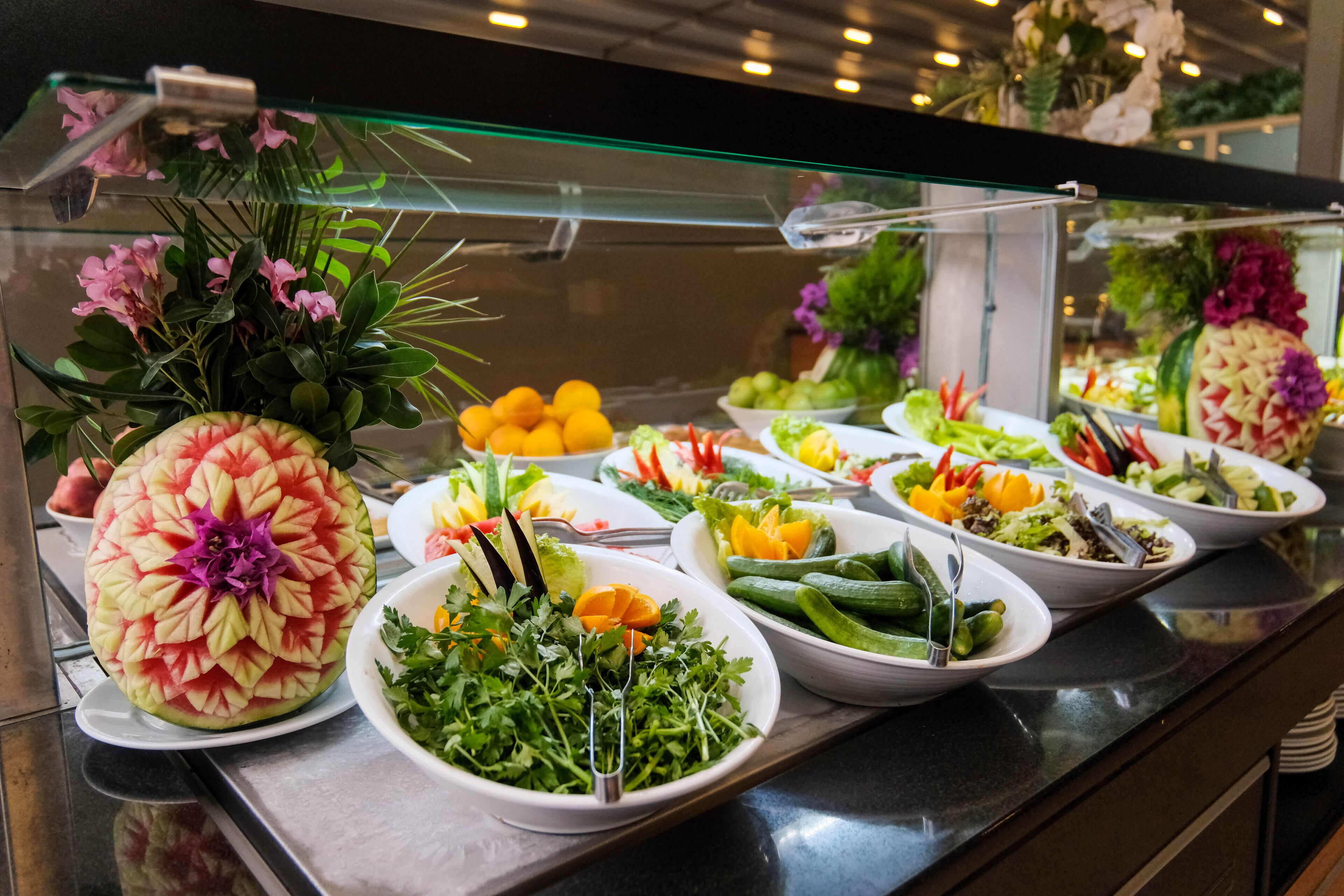 A buffet with various dishes including a watermelon carved into a flower, fresh greens, and assorted vegetables.
