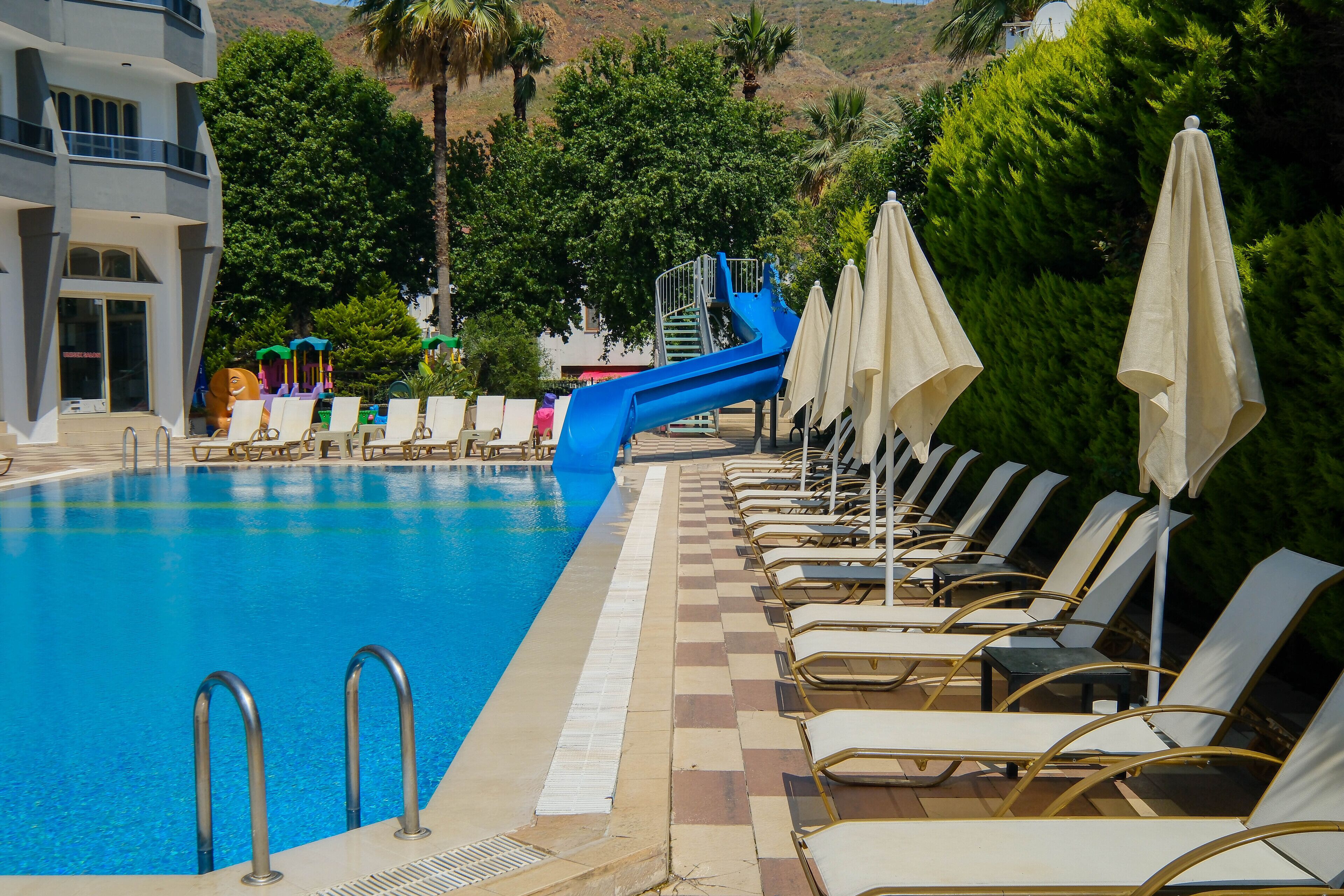A swimming pool with lounge chairs and umbrellas, a blue slide, and a building with a balcony.