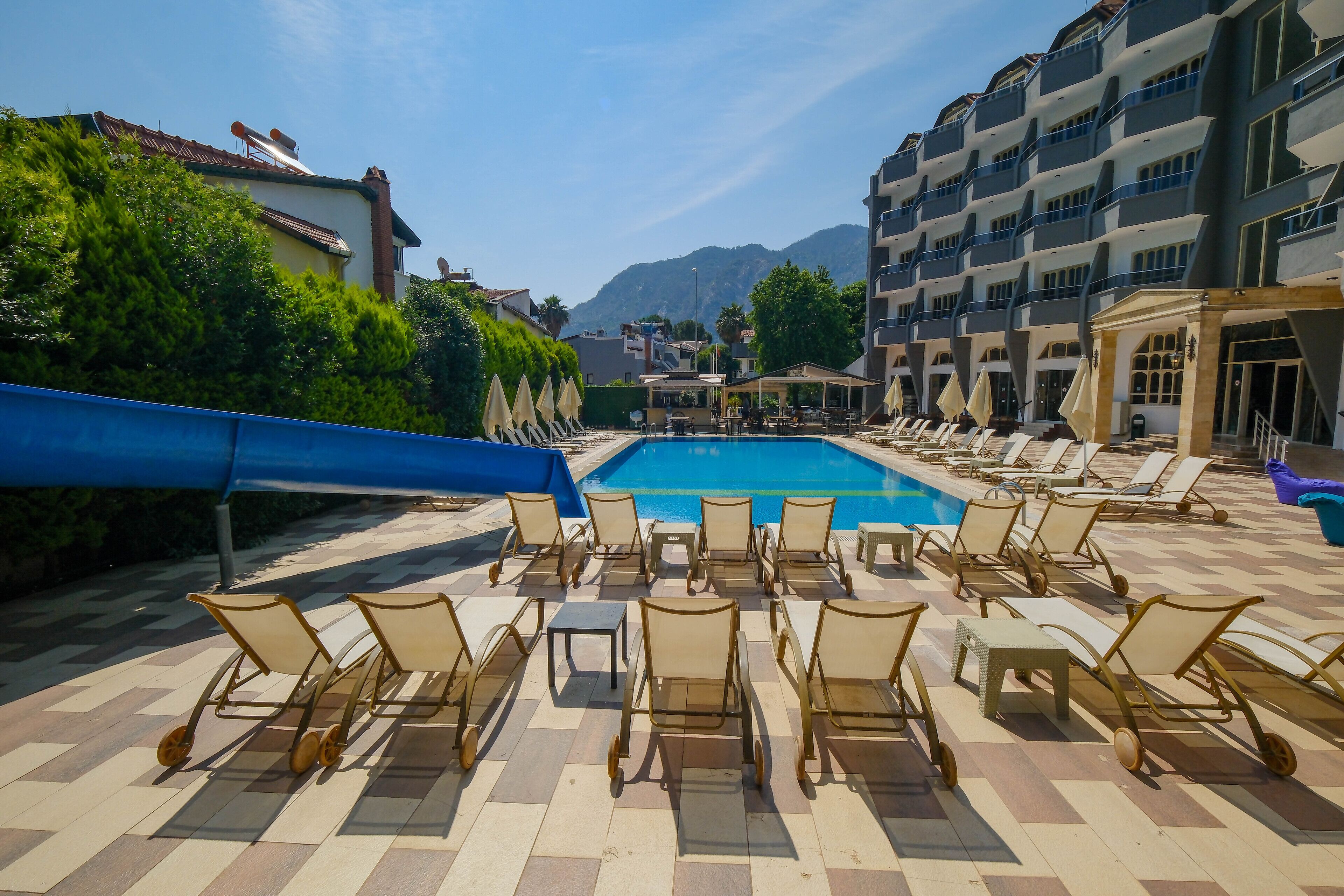 A poolside area with lounge chairs and umbrellas, a swimming pool, and a view of mountains in the background.