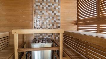Un cuarto de sauna con bancos de madera, pared de azulejos y un recipiente metálico probablemente para agua o vapor.