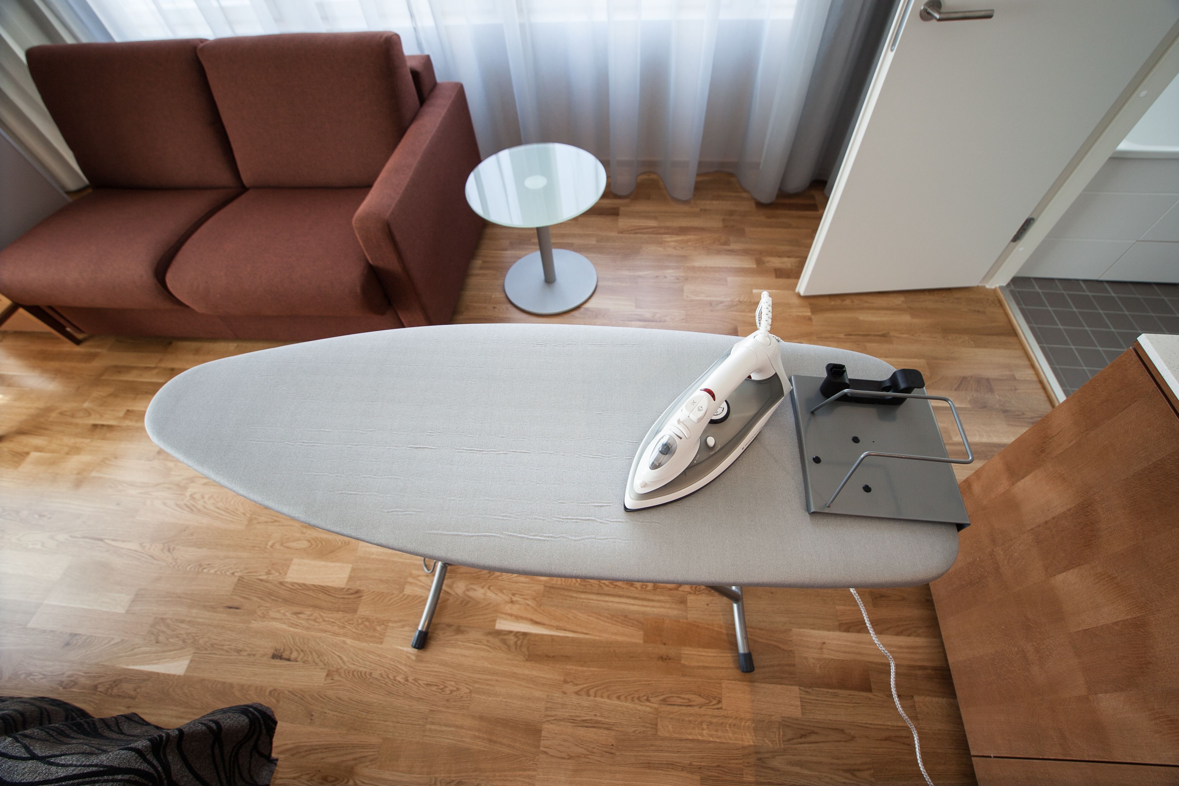 Una habitación moderna con suelo de madera, una tabla de planchar gris, un planchador y un sofá marrón.