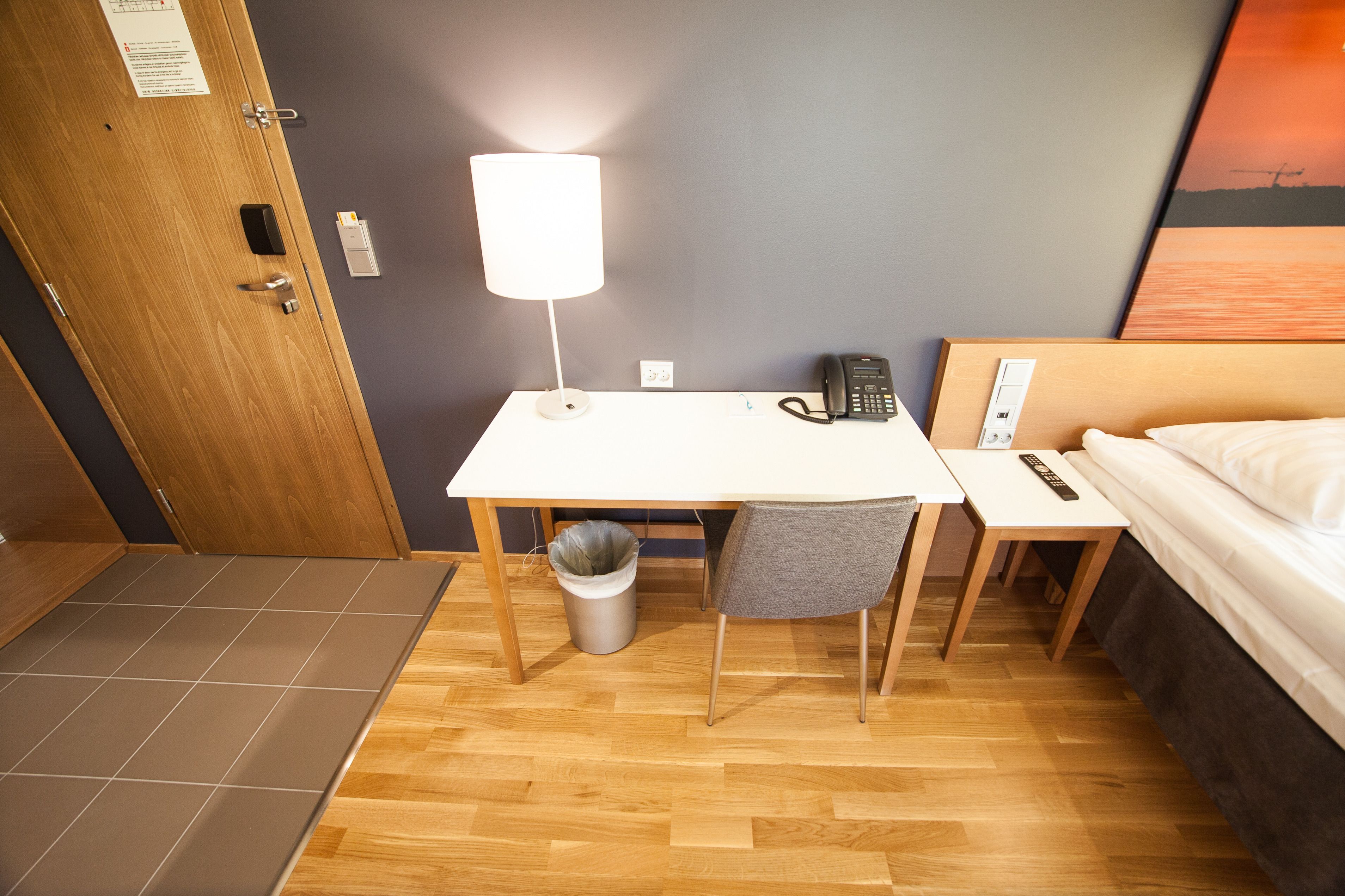 Habitación de hotel con un escritorio de madera, una silla, una cama, una mesita de noche, una lámpara, un teléfono y un cubo de basura.