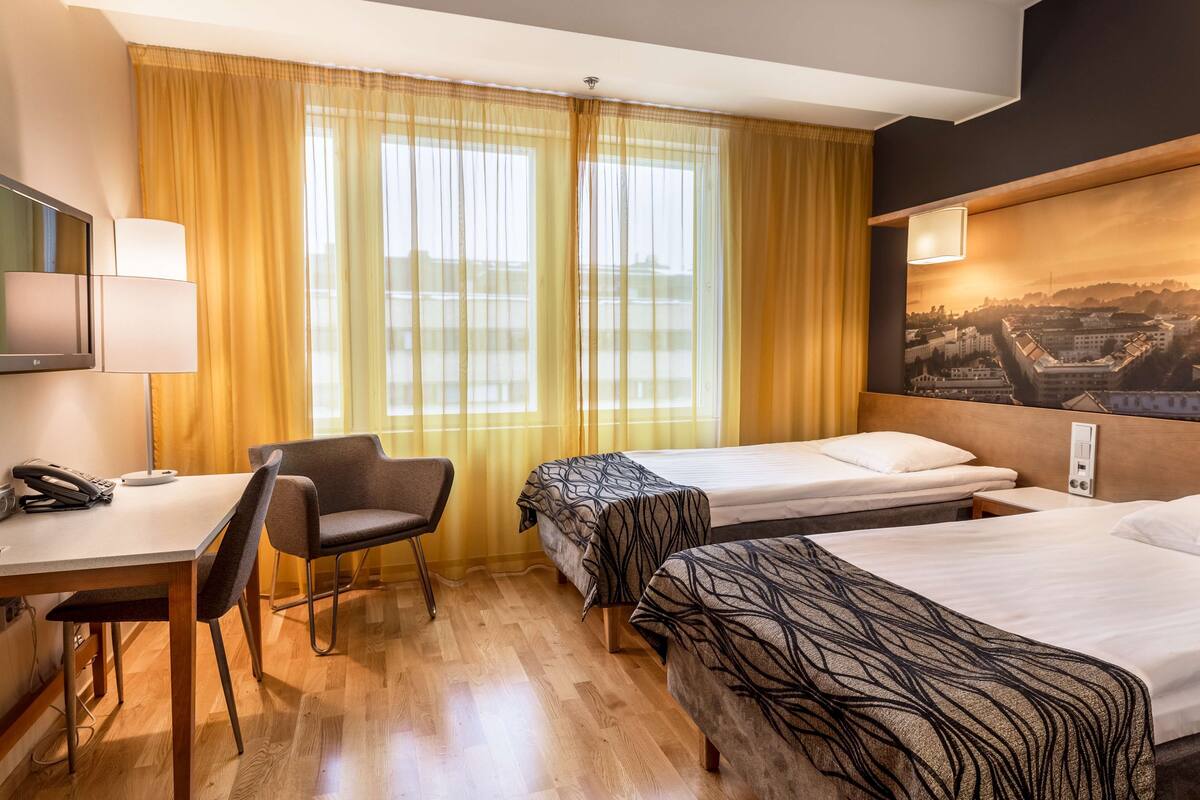 Une chambre d’hôtel avec deux lits, un bureau avec une chaise, une télévision et une fenêtre avec des rideaux.