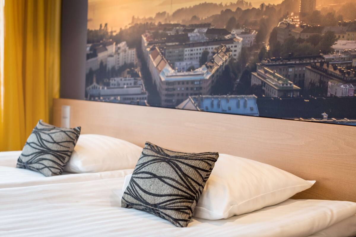 Une chambre d’hôtel avec un lit, deux coussins à motifs et une vue sur la ville par la fenêtre.