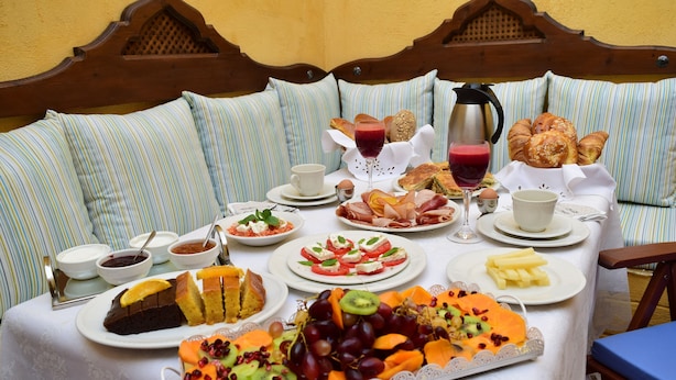 Une table de petit-déjeuner, dressée sur un lit orné d’oreillers à rayures, est garnie de divers plats, notamment des viennoiseries, des fruits et des boissons.