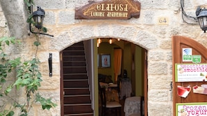 Un bâtiment en pierre avec une enseigne indiquant « ELFOSSI, HÔTEL DE LUXE », et une porte sur laquelle on peut lire les mentions « smoothies », « yaourts glacés » et « fruits frais ».