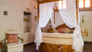 Une chambre à coucher avec un lit à baldaquin en bois, un fauteuil à motifs floraux et une étagère ornée d’objets décoratifs.