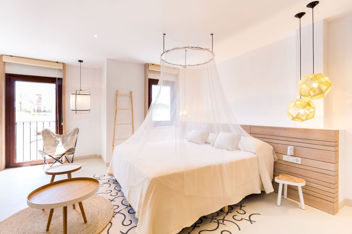 A modern bedroom with a large bed, a wooden headboard, a small stool, a chair, and a balcony view.