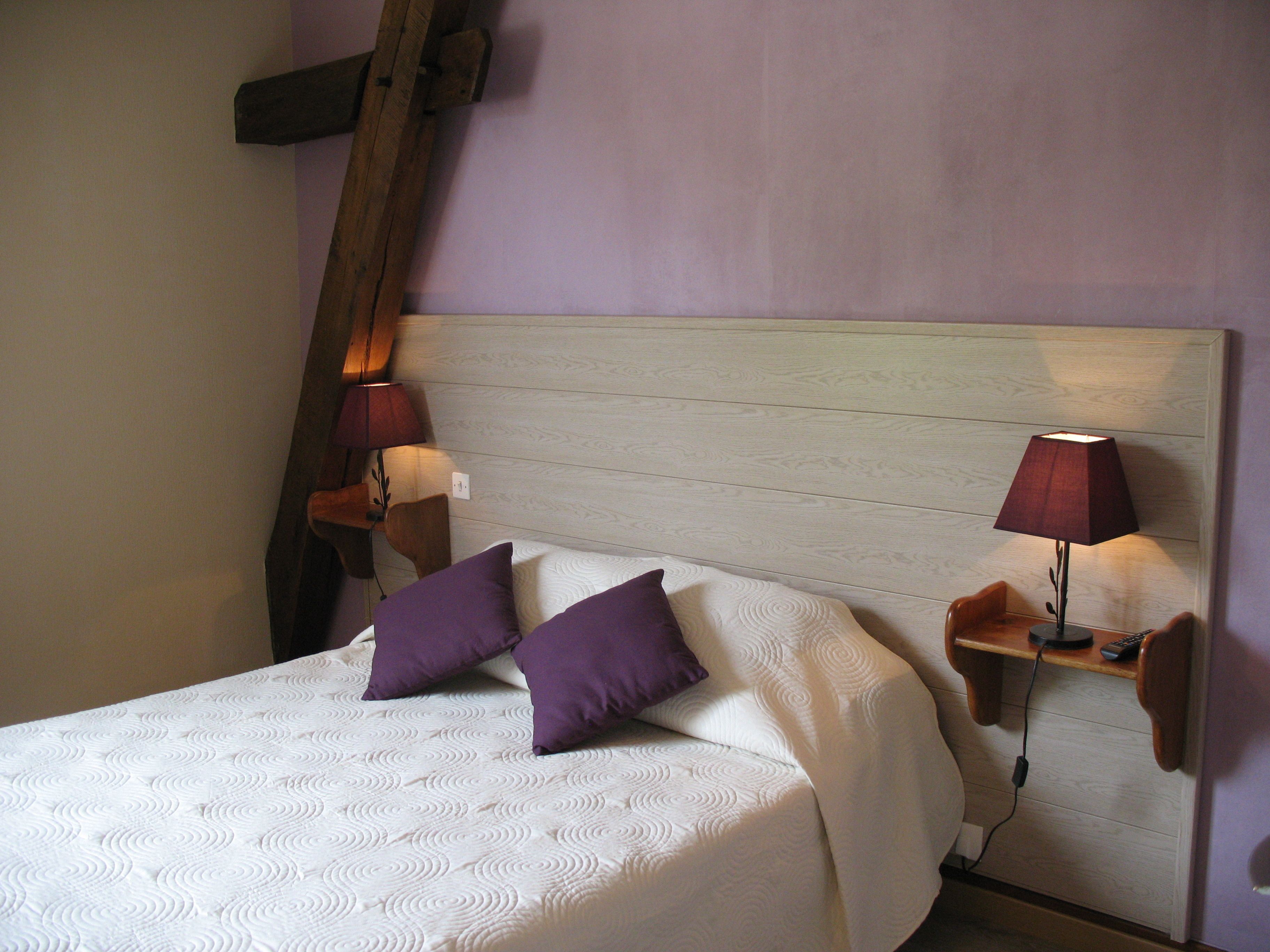 A neatly made bed with a white quilt and two purple pillows, flanked by two bedside tables with lamps, in a room with wooden paneling and a sloped ceiling.