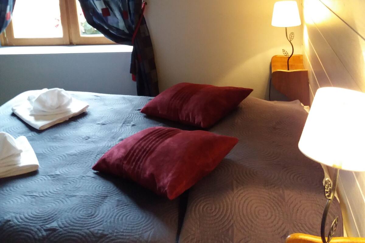 Habitación de hotel con cama, mesita de noche con lámpara, ventana con cortinas y cabecero fijado a la pared.
