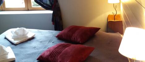 Quarto de hotel com cama, mesa de cabeceira com abajur, janela com cortinas e cabeceira fixada na parede.