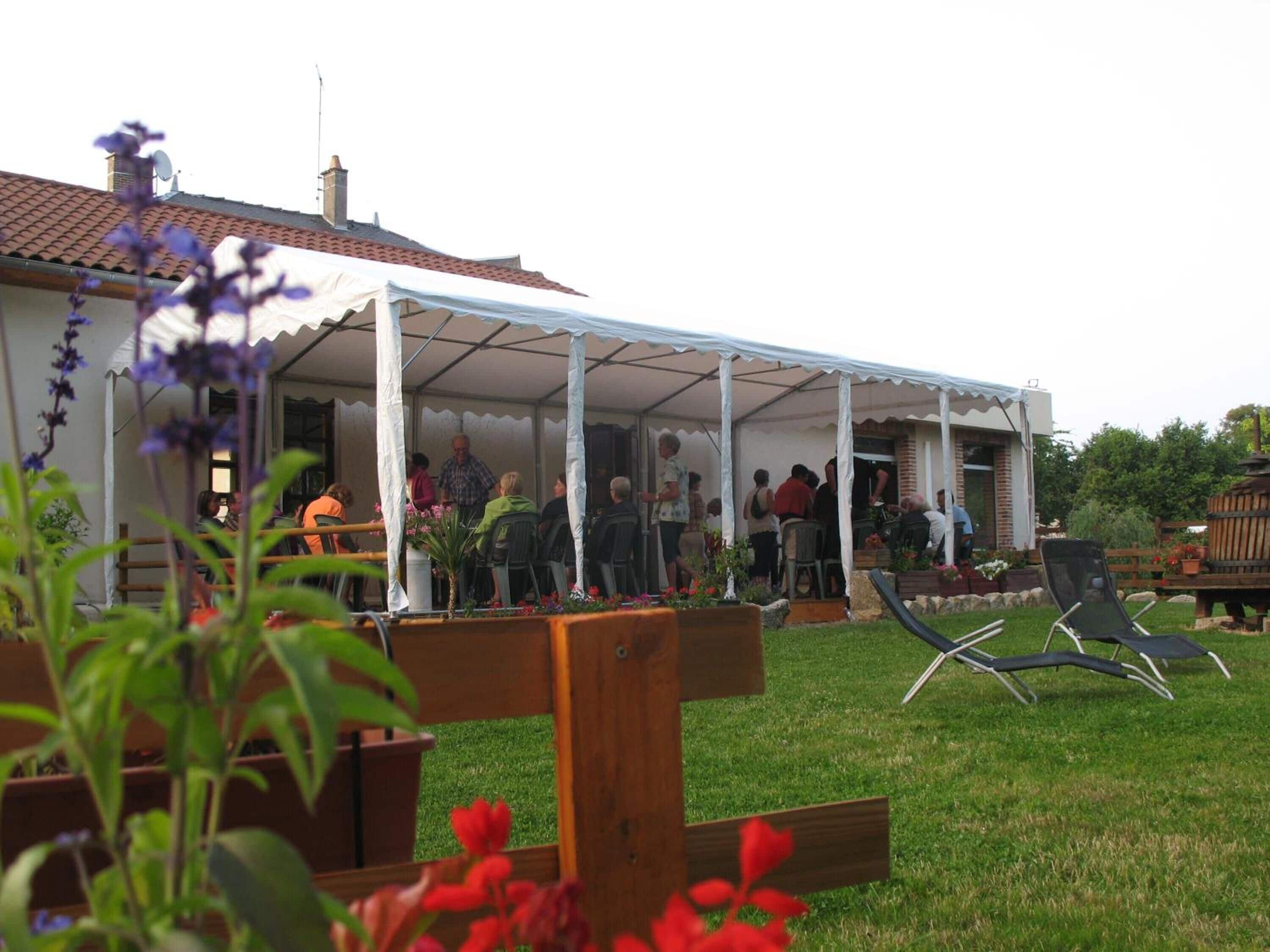 A covered outdoor seating area with people gathered around tables and chairs, a grassy lawn, and a building with a tiled roof in the background.