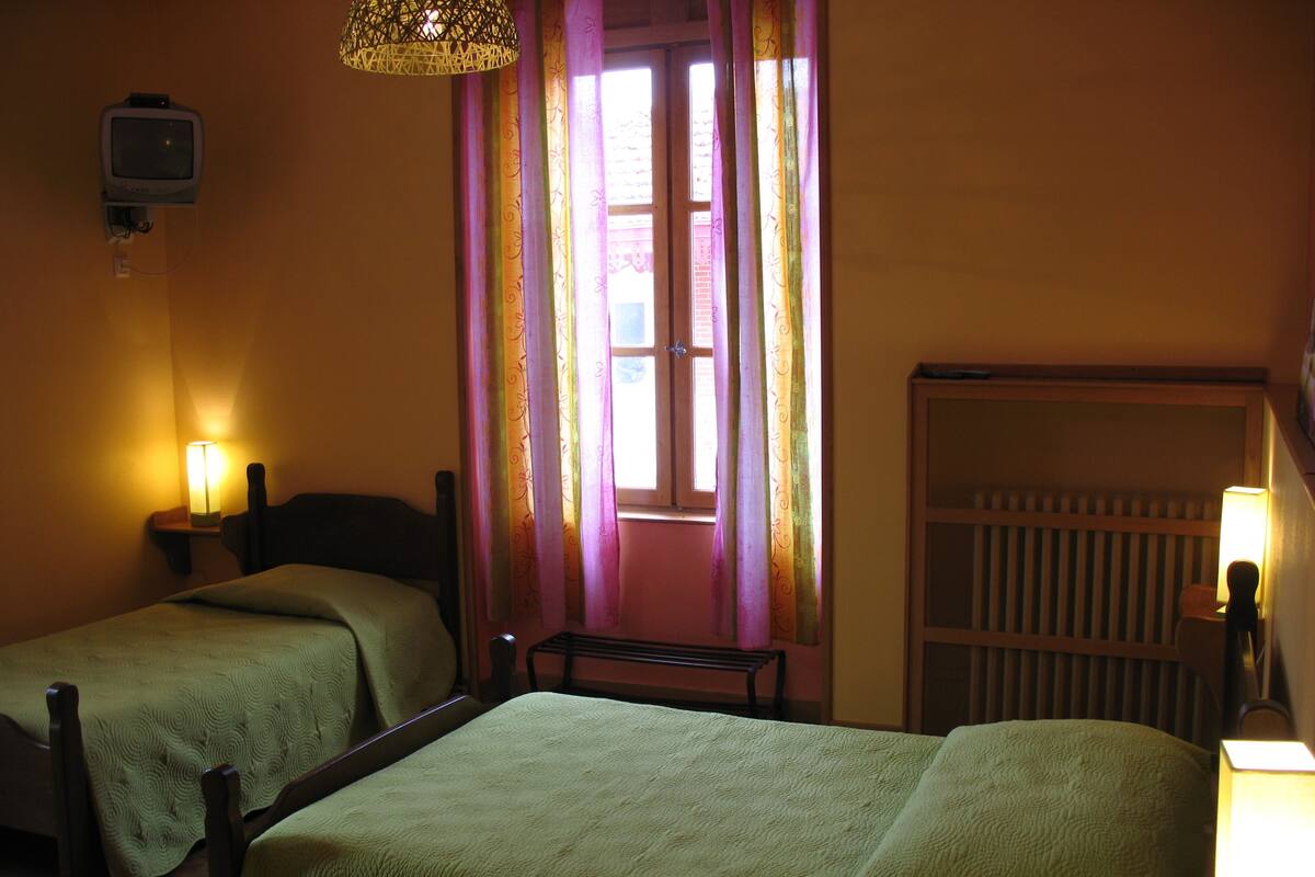 Una habitación con dos camas individuales, una mesita pequeña, televisión instalada en la pared y una ventana con cortinas.