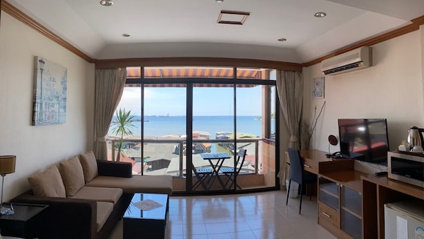 A modern living room with a large window overlooking the sea, a sofa, a coffee table, a TV, and a dining area with a table and chairs. - Seaview Sriracha Hotel (Si Racha)