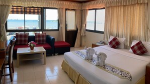 A hotel room with a bed, a sofa, a small table with a vase, and a view of the sea through large windows. - Seaview Sriracha Hotel (Si Racha)