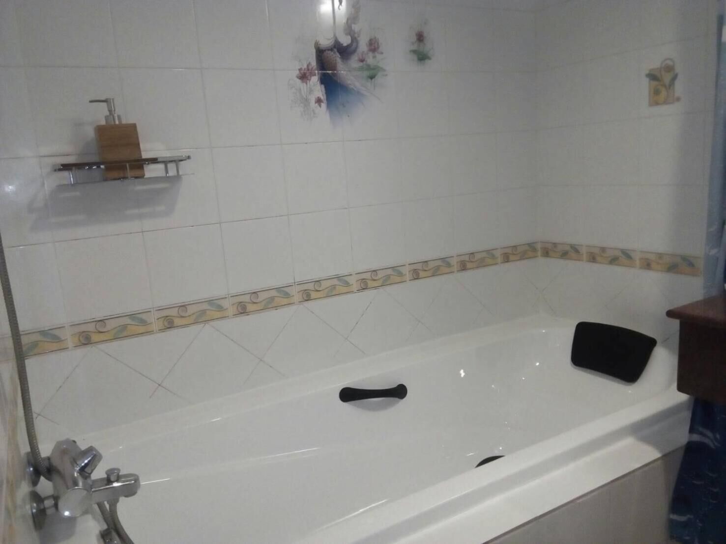 Une salle de bain avec une baignoire, une douche, un distributeur de savon posé sur une étagère et des carreaux décoratifs sur le mur.