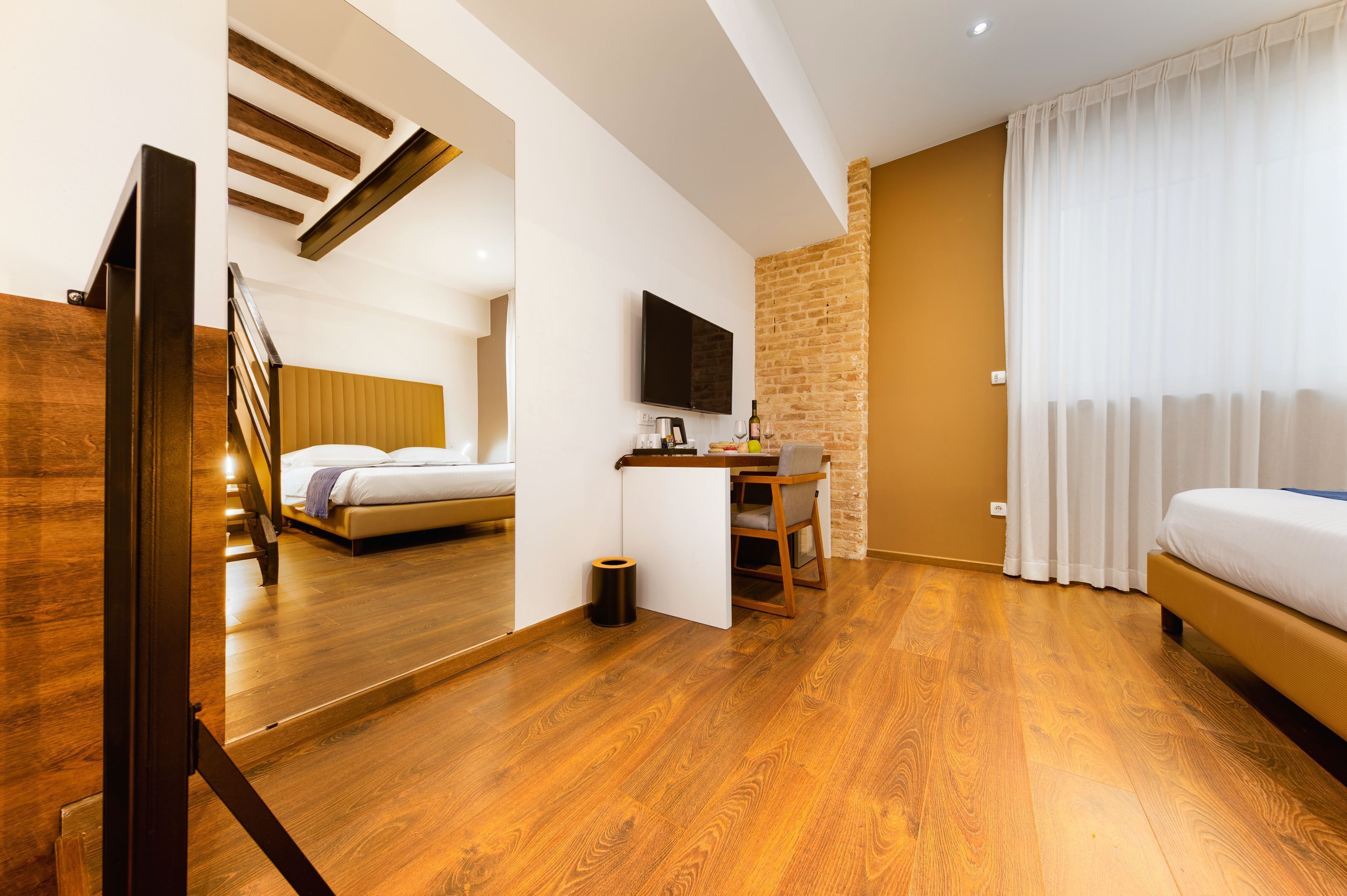 A modern hotel room with wooden flooring, a bed, a desk with a chair, a television, and a brick accent wall.