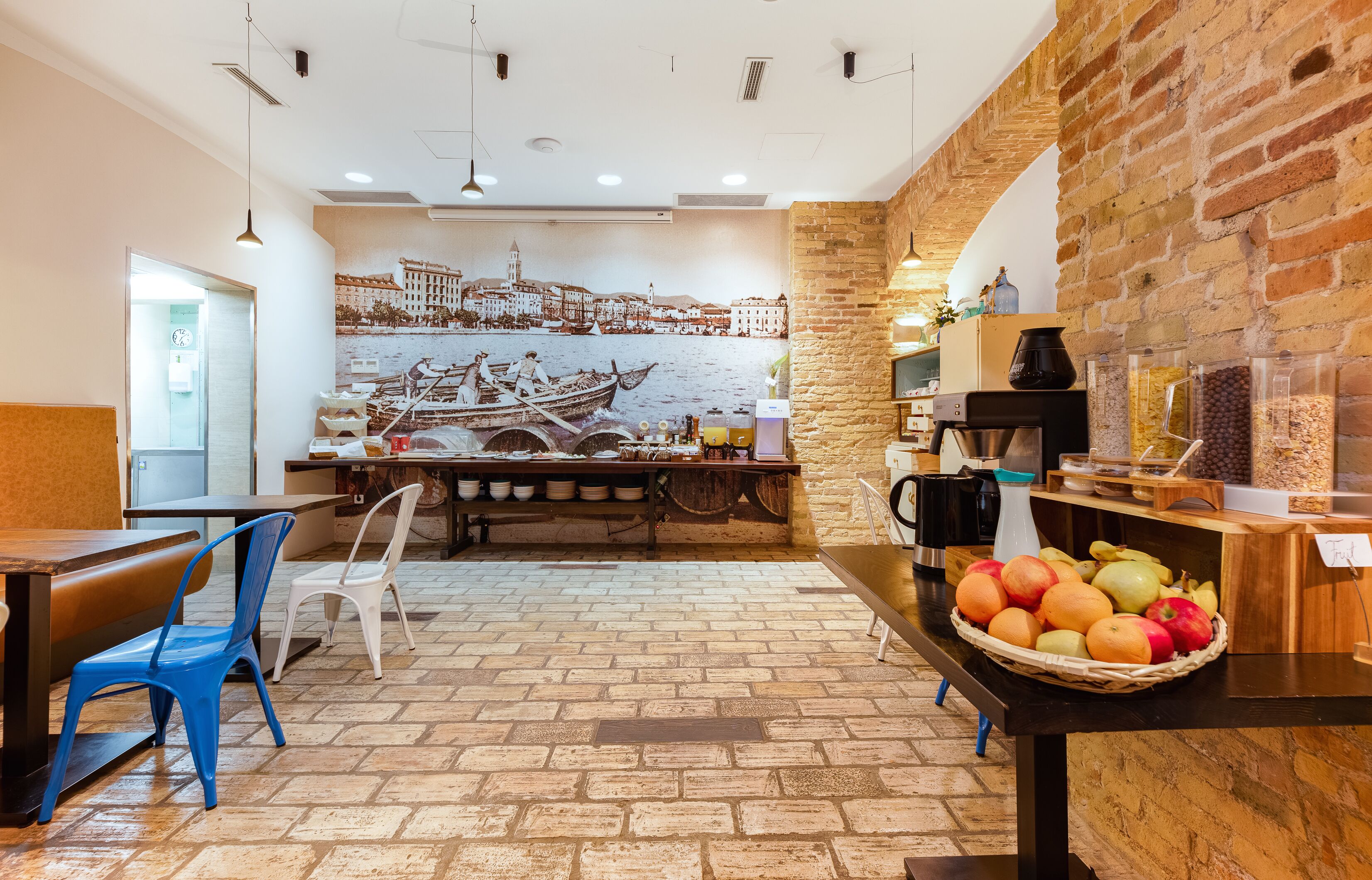 A restaurant with a brick wall, a mural of a coastal town, a buffet counter with fruits, and a seating area with chairs and tables.