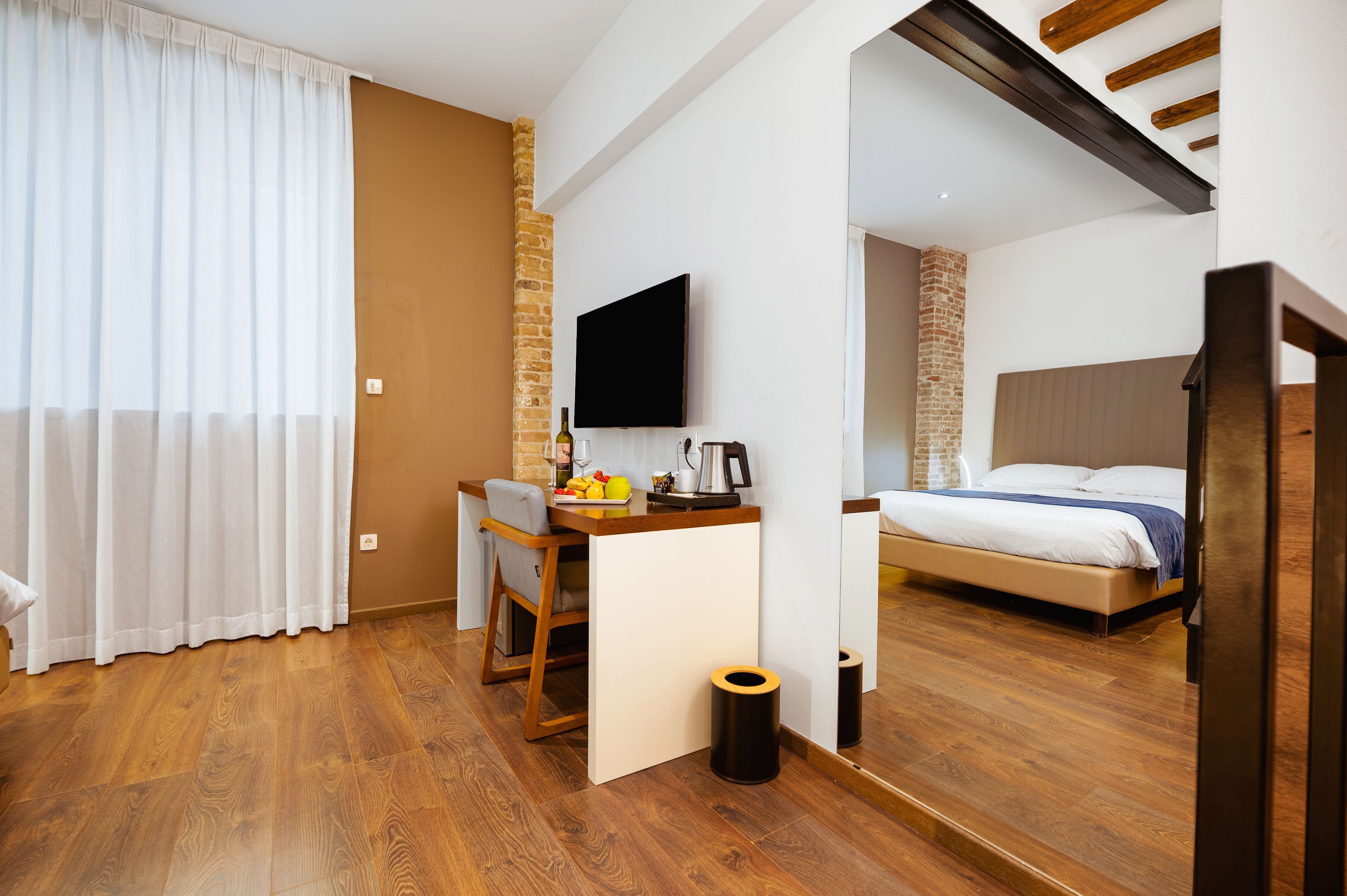 A modern hotel room with a wooden floor, a desk with a chair, a bed, a television, and a brick accent wall.