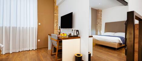 A modern hotel room with a wooden floor, a desk with a chair, a bed, a television, and a brick accent wall.